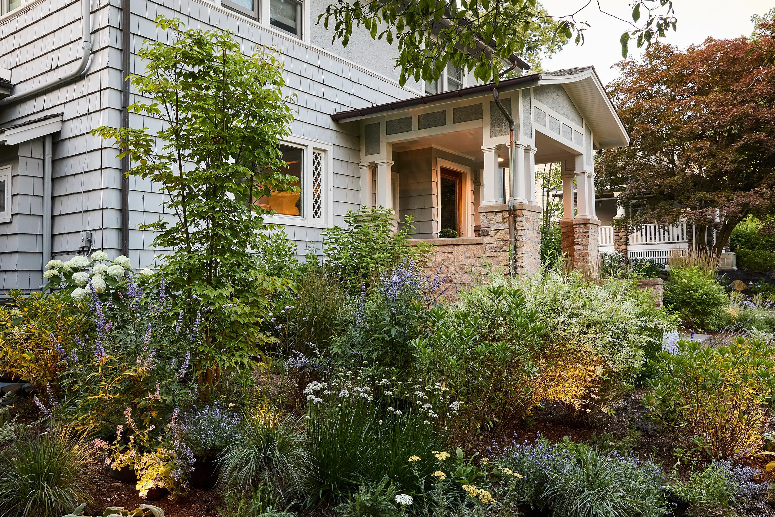 A house with a garden front yard filled with various green plants, flowers, and bushes, a small porch with columns, and exterior walls with shingle siding.