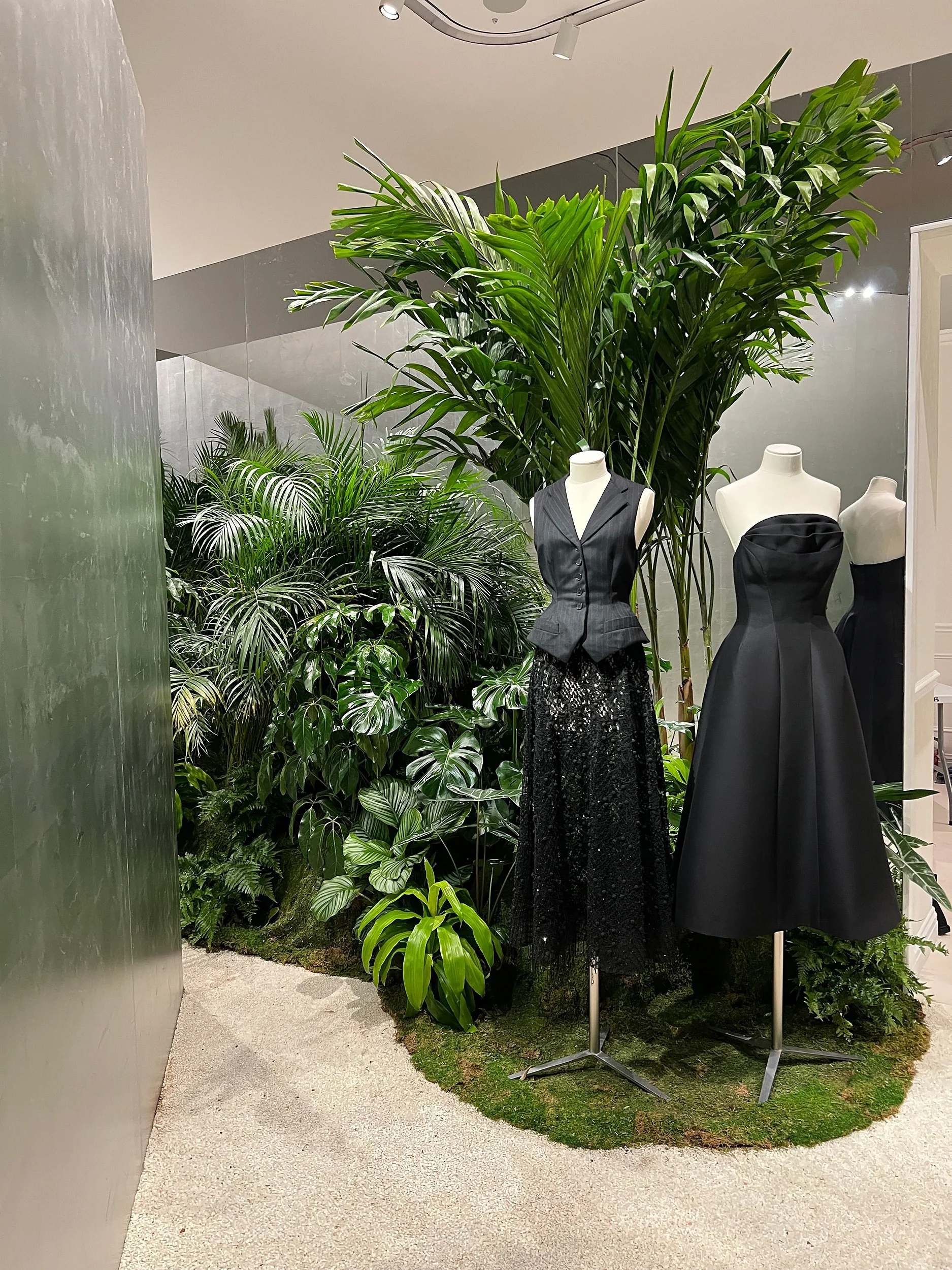Display of four mannequins dressed in black clothing, surrounded by lush green tropical plants in an indoor setting.