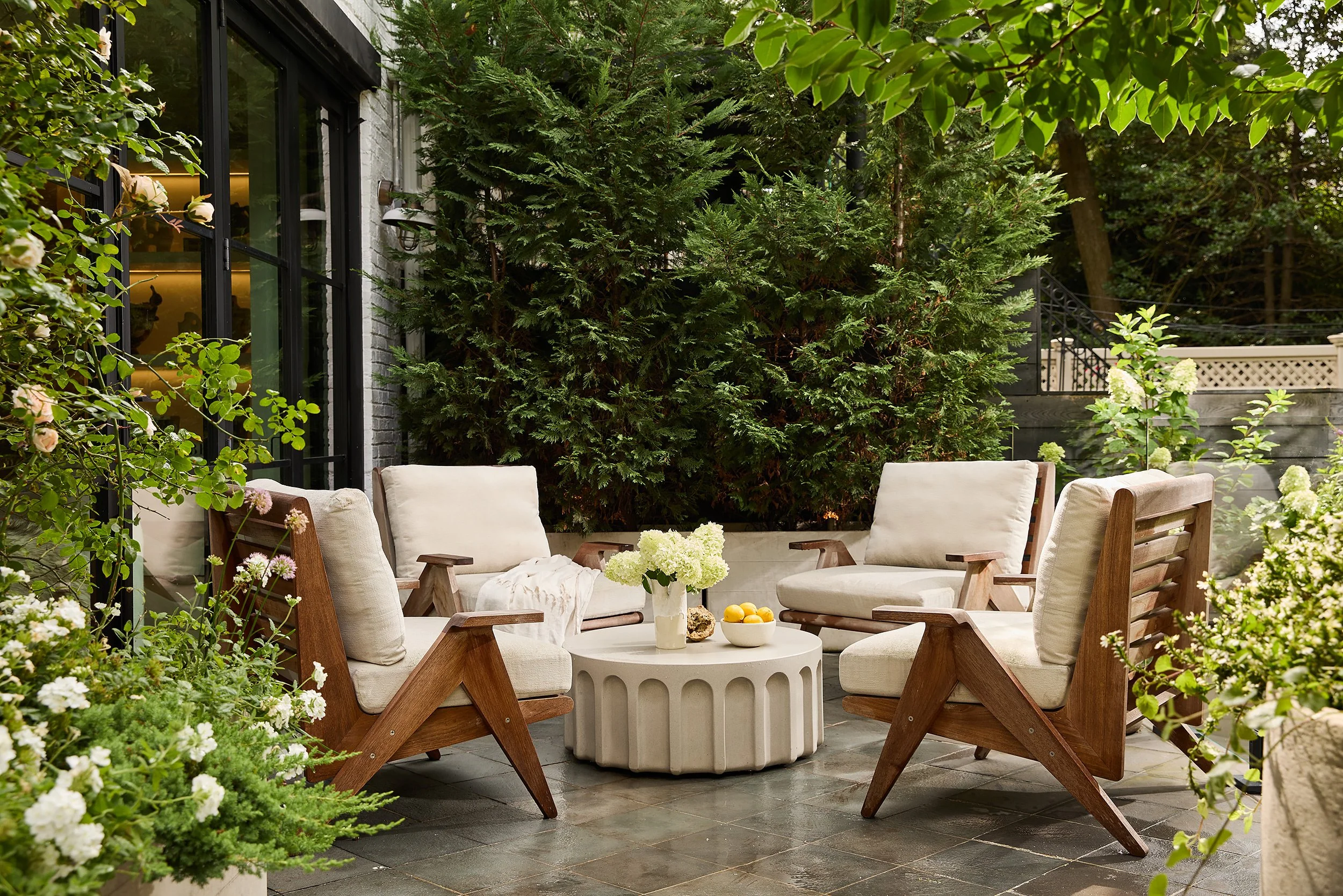 Outdoor patio with four wooden framed chairs with white cushions, arranged around a white round table with a flower vase and bowls of lemons. Surrounded by lush green plants and trees.