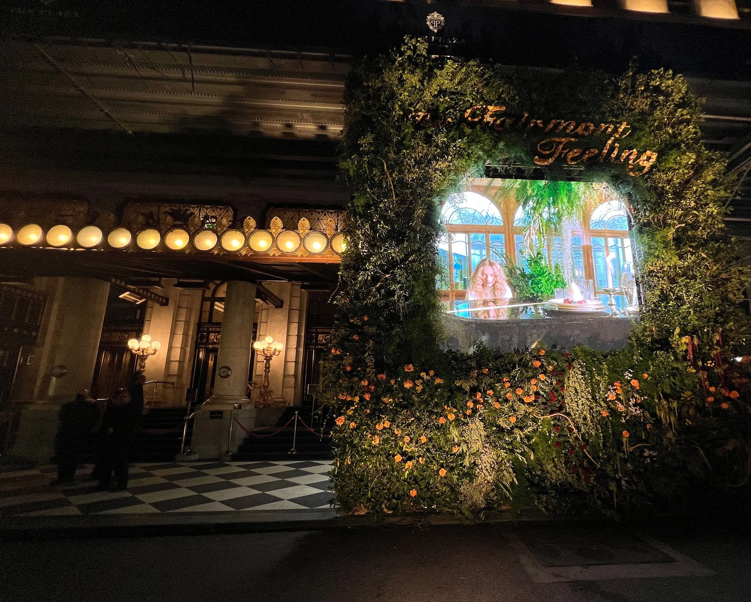 A decorated entrance with a large, illuminated sign that reads 'Momenti Feeling,' surrounded by greenery and flowers. There is a large screen displaying a woman with blonde hair and a bright background of large windows and plants. The setting appears to be an indoor event space or hotel lobby with elegant lighting, columns, and a checkered floor.