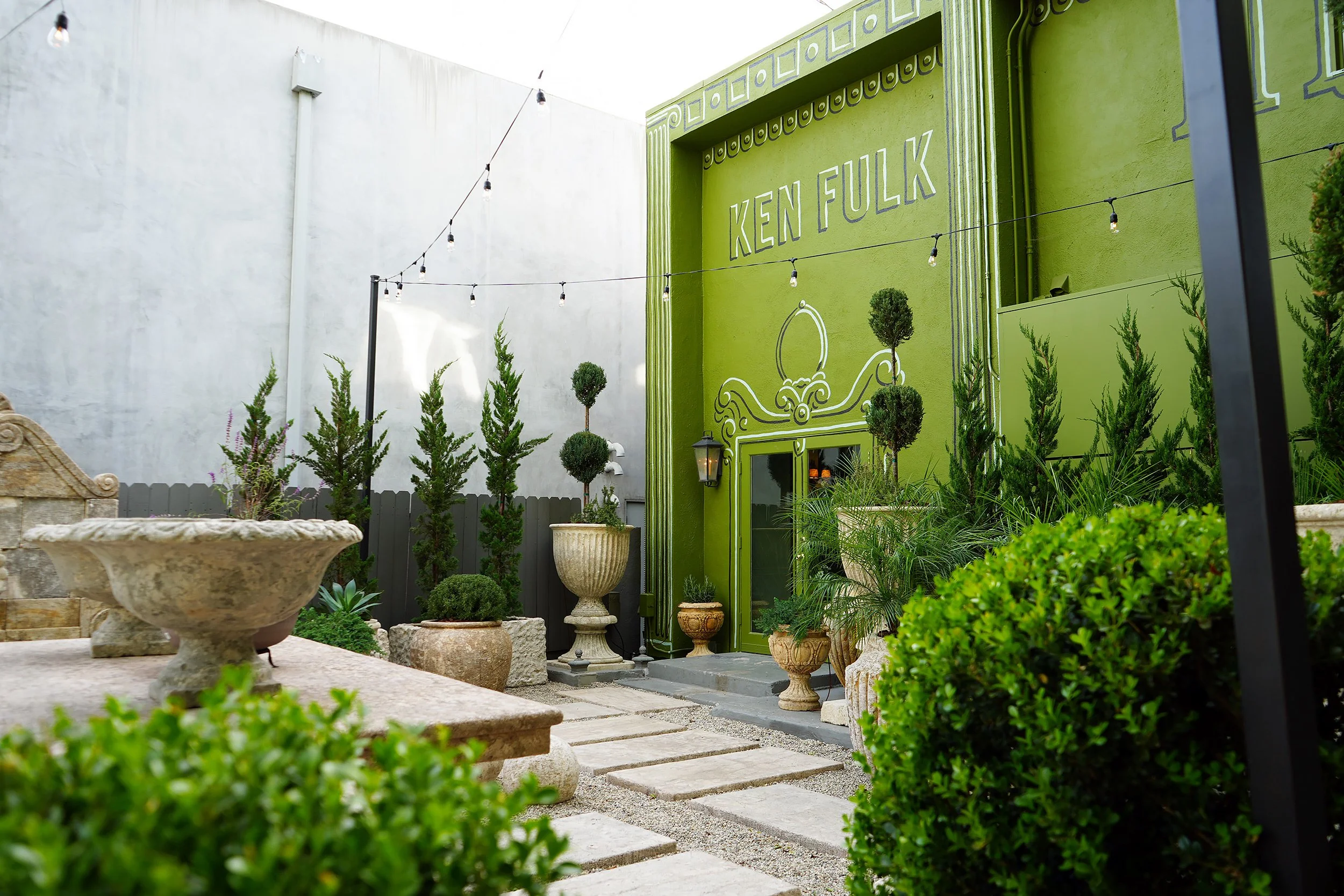 A colorful outdoor patio featuring a bright green wall with the name "Ken Fulks" painted on it and decorated with plants, potted shrubs, and string lights.