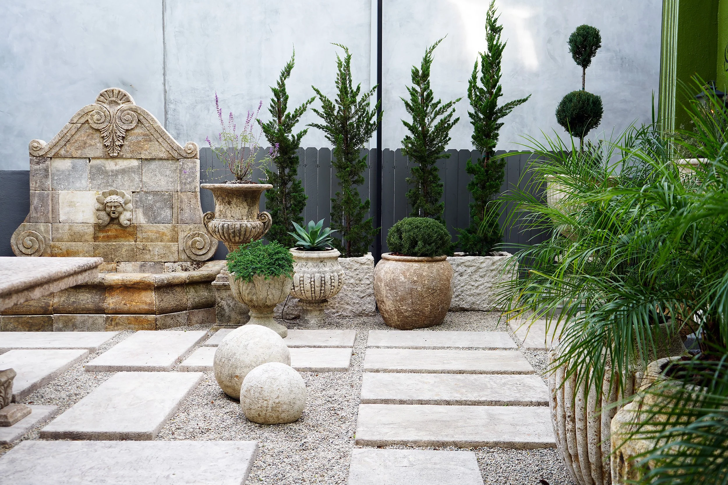 A small outdoor garden with potted plants, a stone fountain, decorative stones, and a gravel pathway, enclosed by a green fence and gray wall.