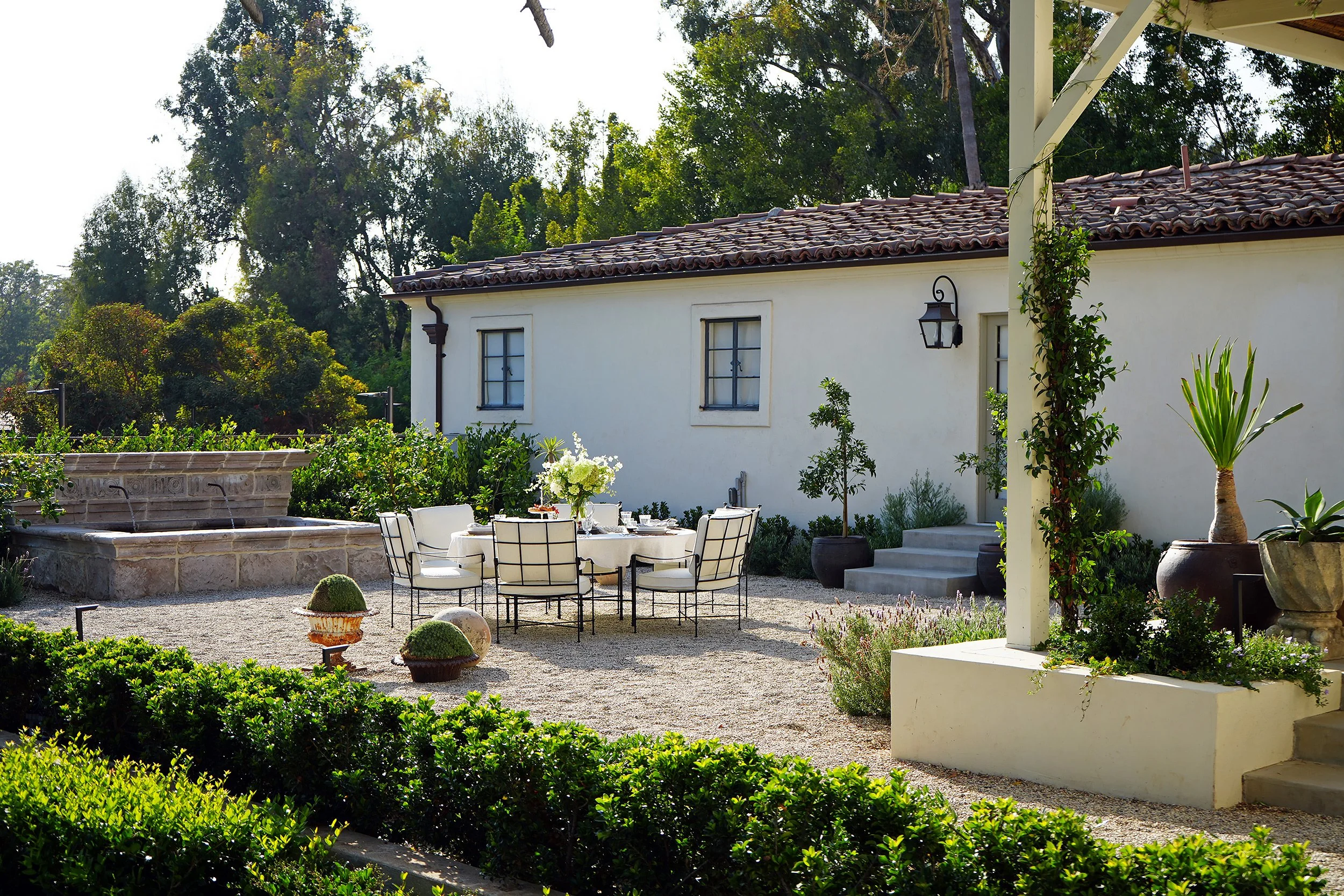 A backyard patio with a white house, outdoor dining table with white chairs, a fountain, potted plants, and lush greenery in the background.