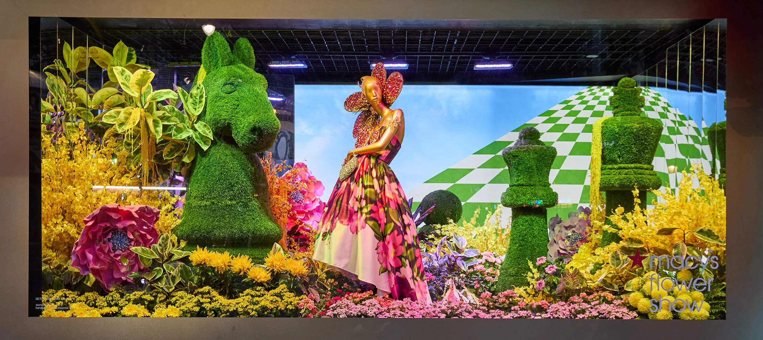 Decorative window display for Macy's Flower Show featuring topiary animals, a mannequin in a floral dress, and a colorful garden scene with large flowers, plants, and a checkered hill in the background.
