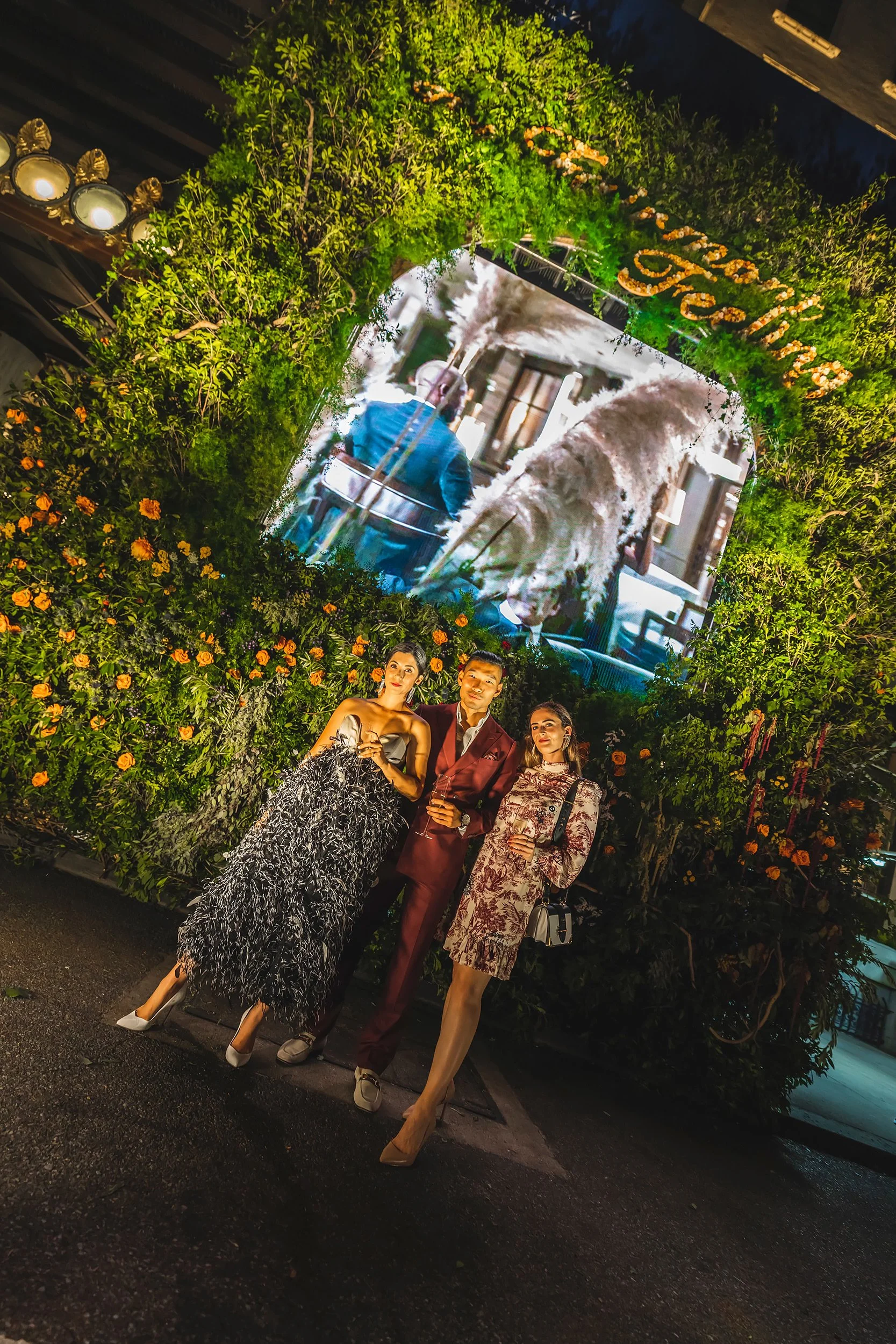 Three well-dressed people standing in front of a decorative wall with green foliage and orange flowers at night. Behind them is a large outdoor LED screen displaying a scene with a lion and a person in a blue jacket.
