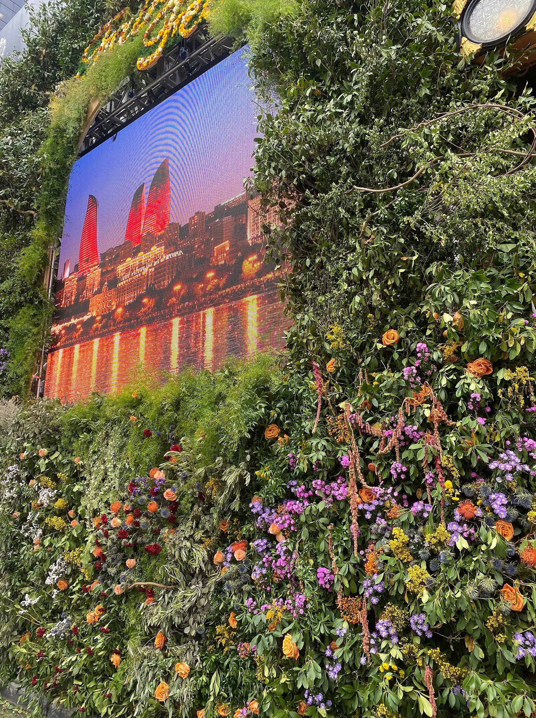 Large digital screen displaying a city skyline at sunset, framed by lush green plants and colorful flowers.