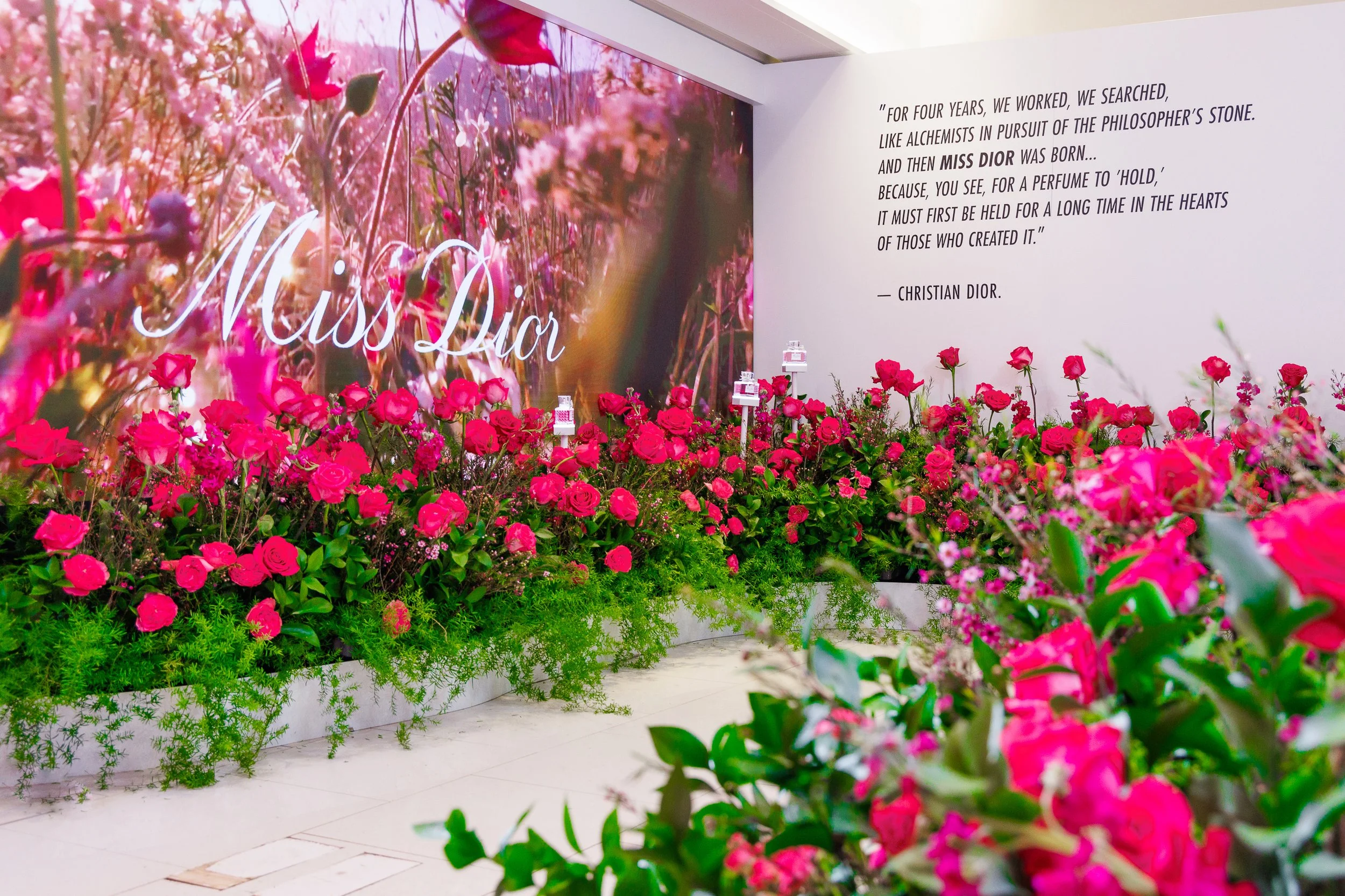 Indoor display of pink roses and greenery at a Miss Dior perfume event, with a large floral-themed banner, Christian Dior quote on the wall, and perfume bottles on stands.