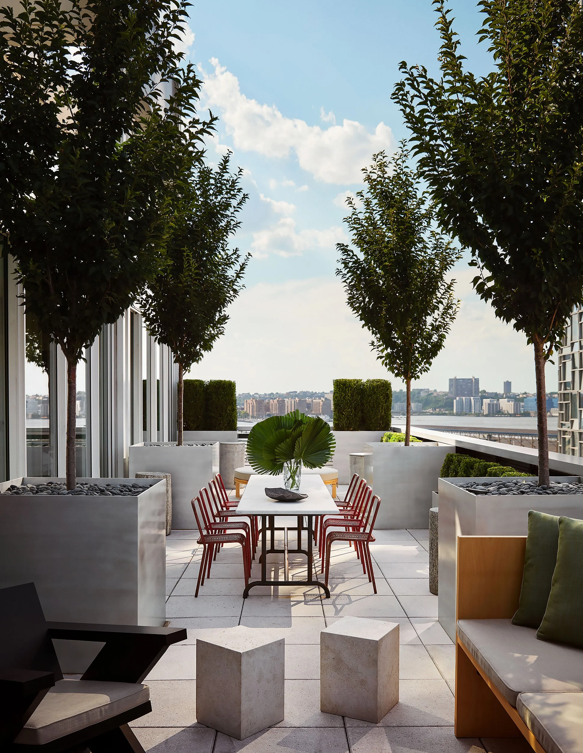 A rooftop outdoor patio with a modern design, featuring a long white table with red chairs, potted trees and plants in white planters, and a view of a city skyline across the water.