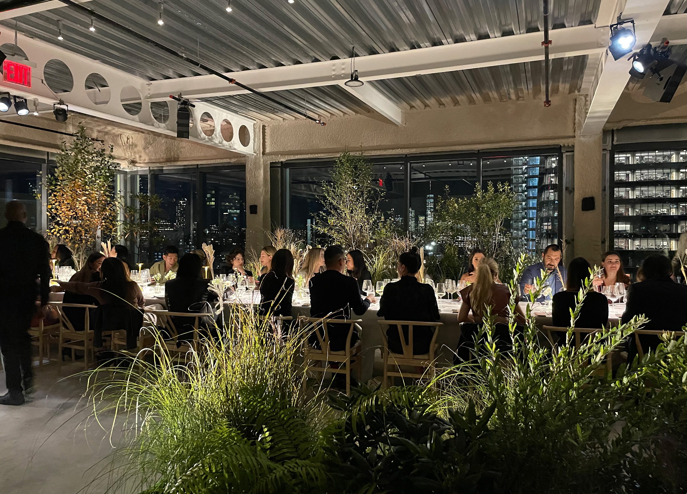 People dining at a long table during a dinner event in a modern, urban restaurant with large windows, city lights visible outside, and lush indoor plants.