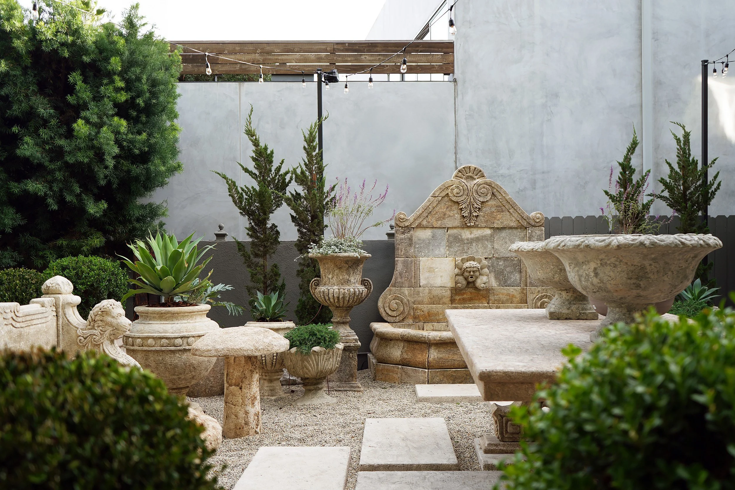 A small, private outdoor patio decorated with classical stone garden ornaments including urns, a fountain, and a bench, surrounded by lush greenery and string lights.