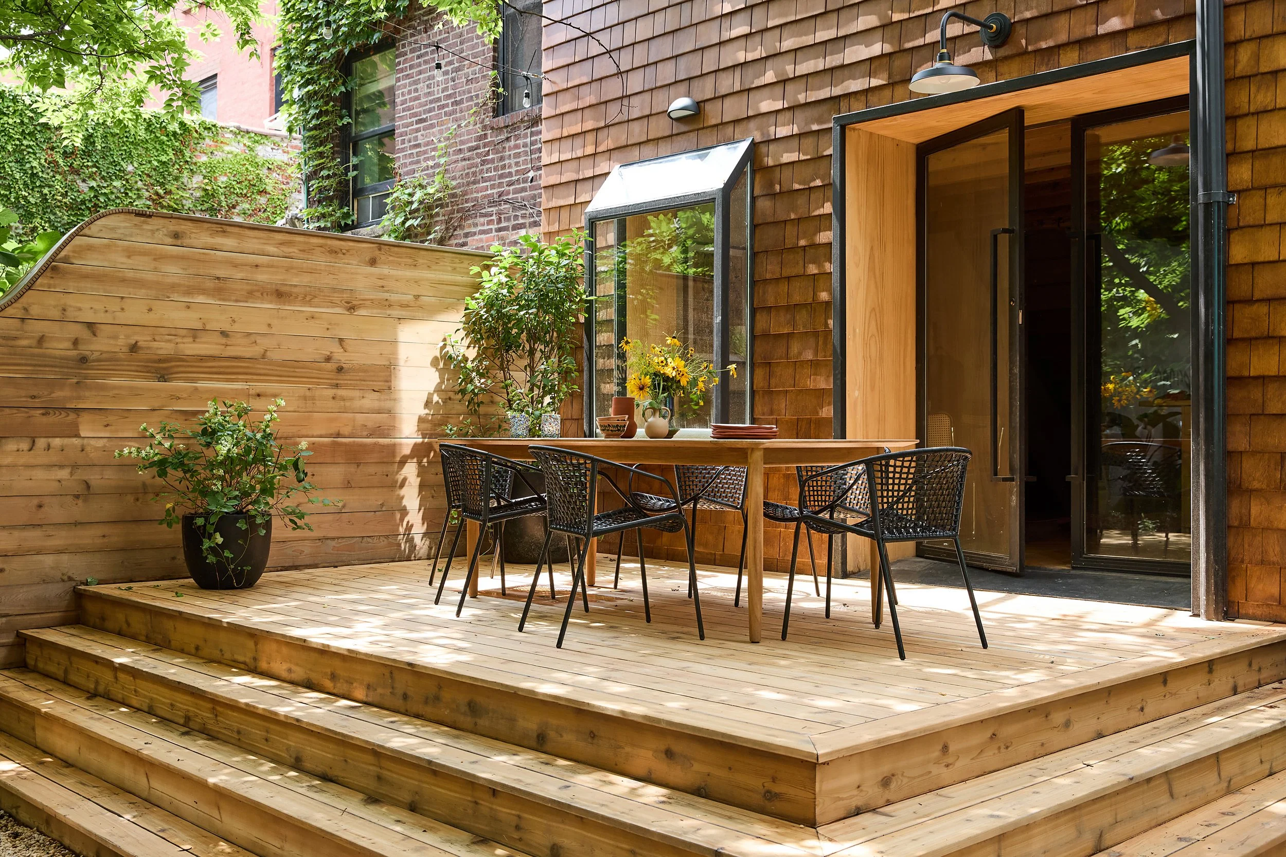 Wooden patio with a round table, six black chairs, potted plants, and a sliding glass door leading inside a brick and wooden house.