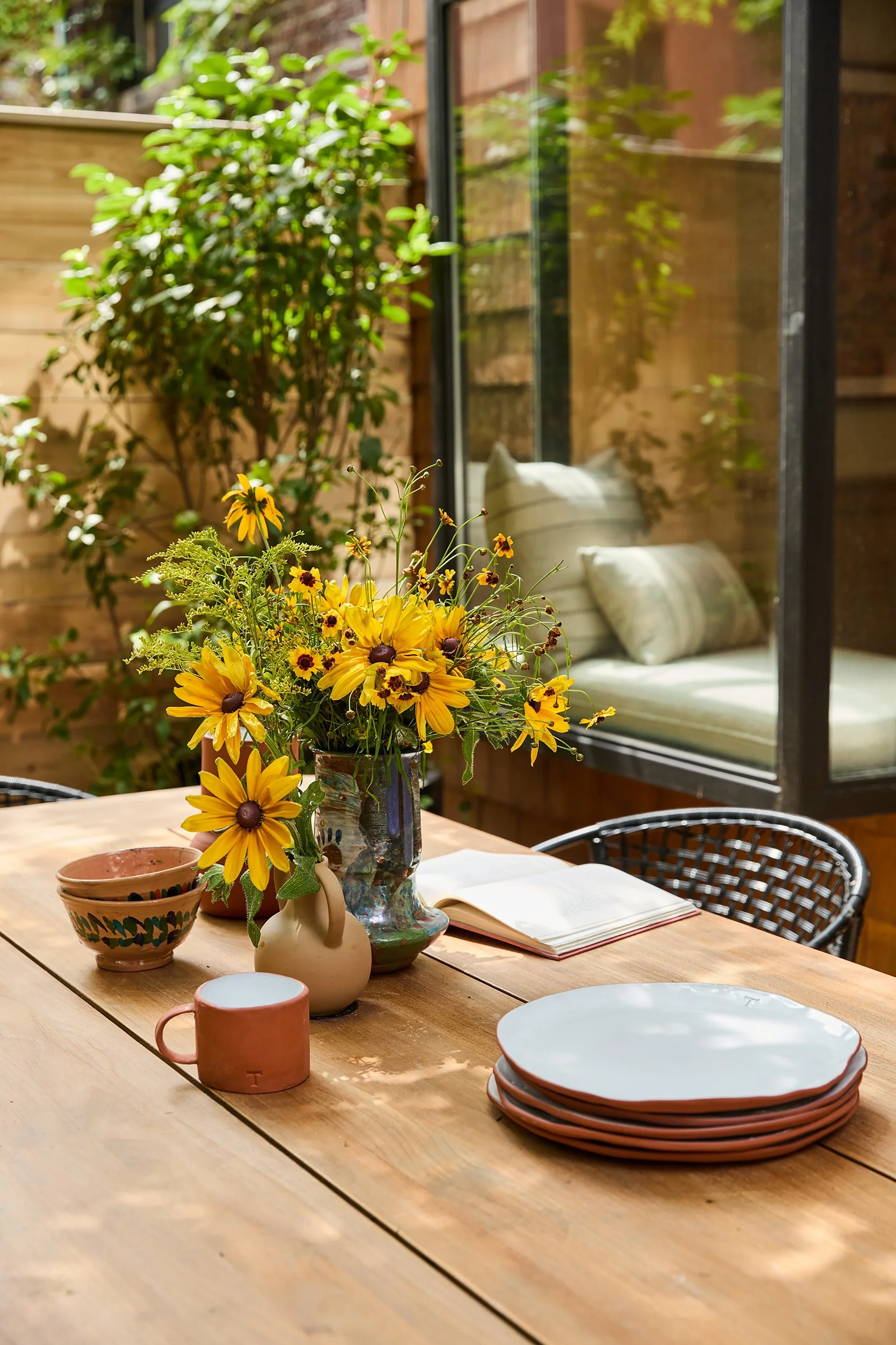 A wooden dining table with a vase of yellow flowers, ceramic bowls, a pink mug, an open book, and stacked white plates. In the background, there's a sitting area with cushions, and a glass window revealing a green outdoor background with plants.