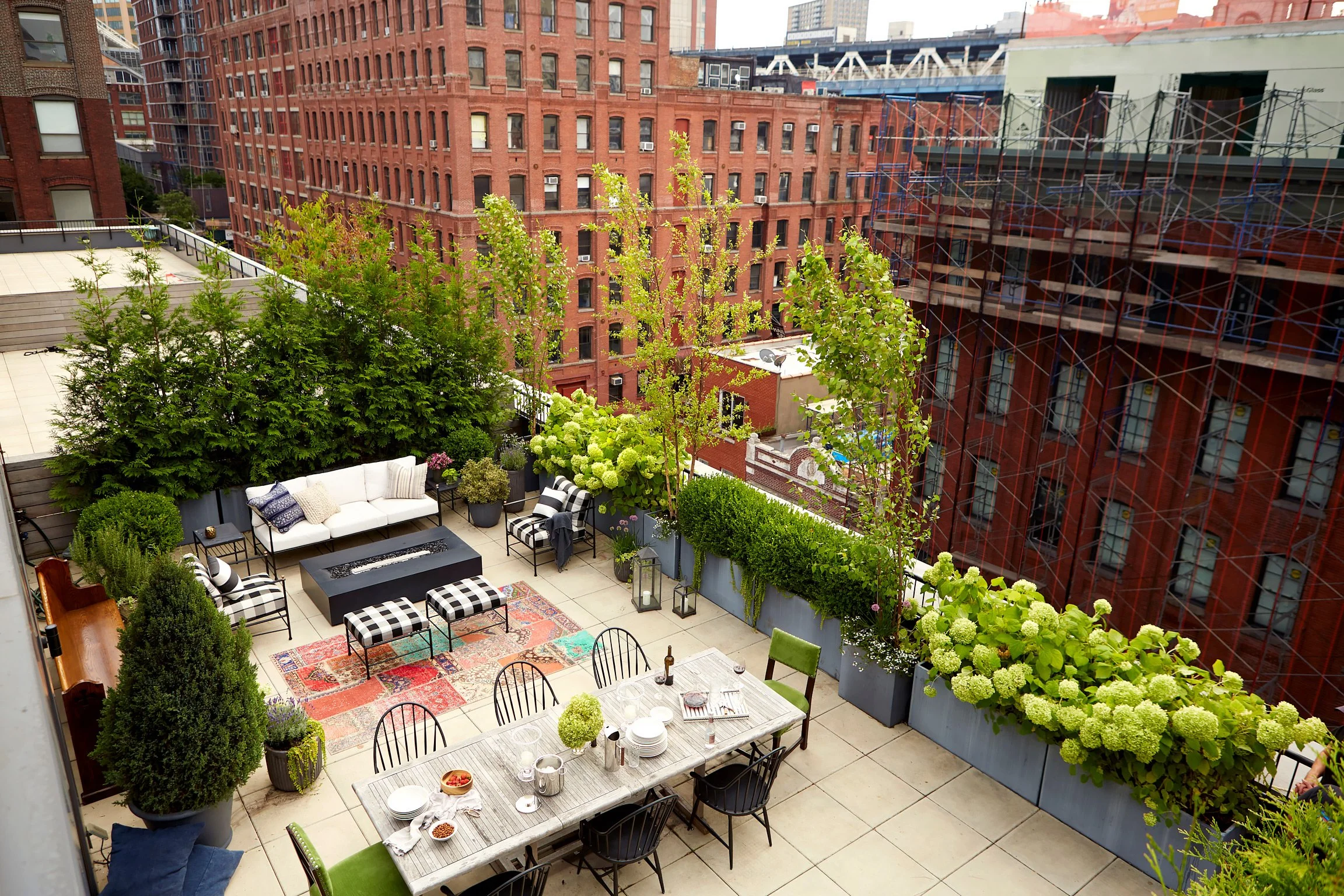City rooftop terrace with outdoor furniture, potted plants, trees, and a dining table set for a meal, surrounded by tall brick buildings and construction scaffolding.