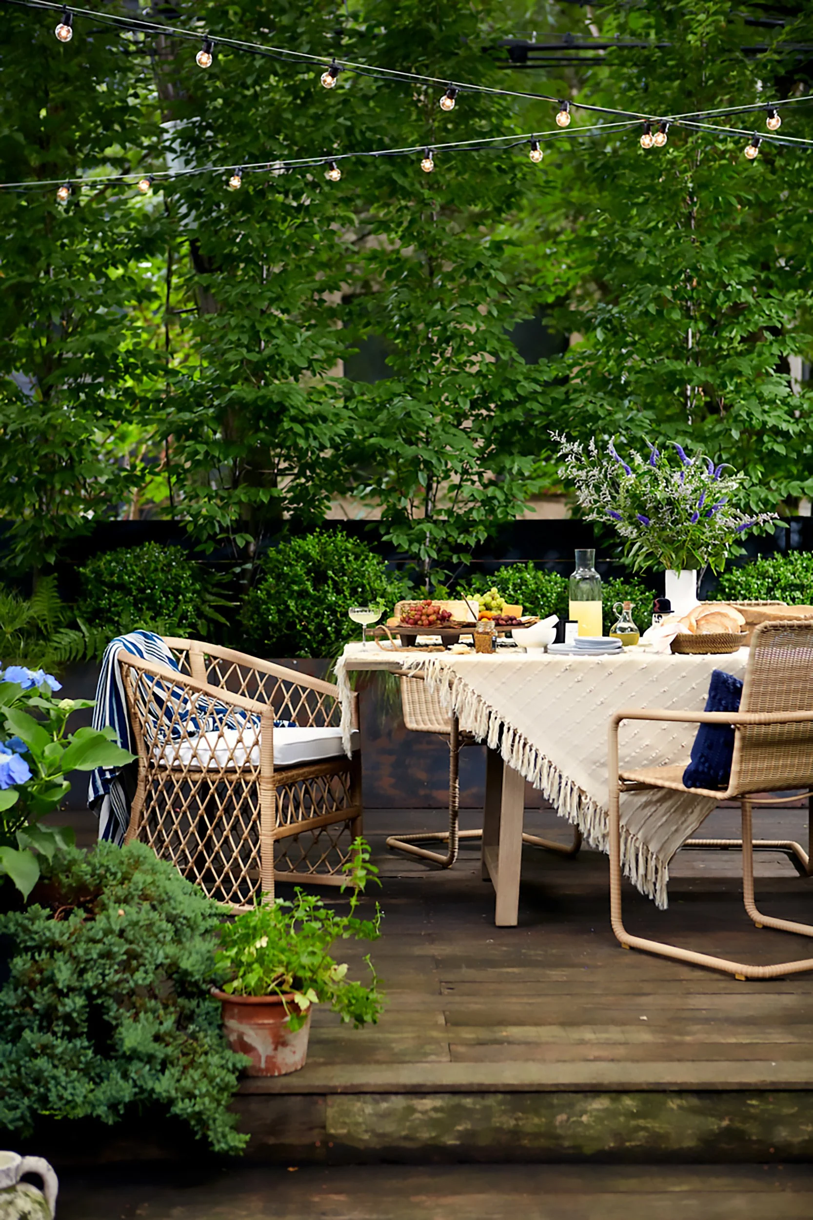 An outdoor dining table set on a wooden deck, decorated with a cream-colored tablecloth, surrounded by wicker chairs, with a large flower arrangement and food and drinks, under string lights and lush greenery.