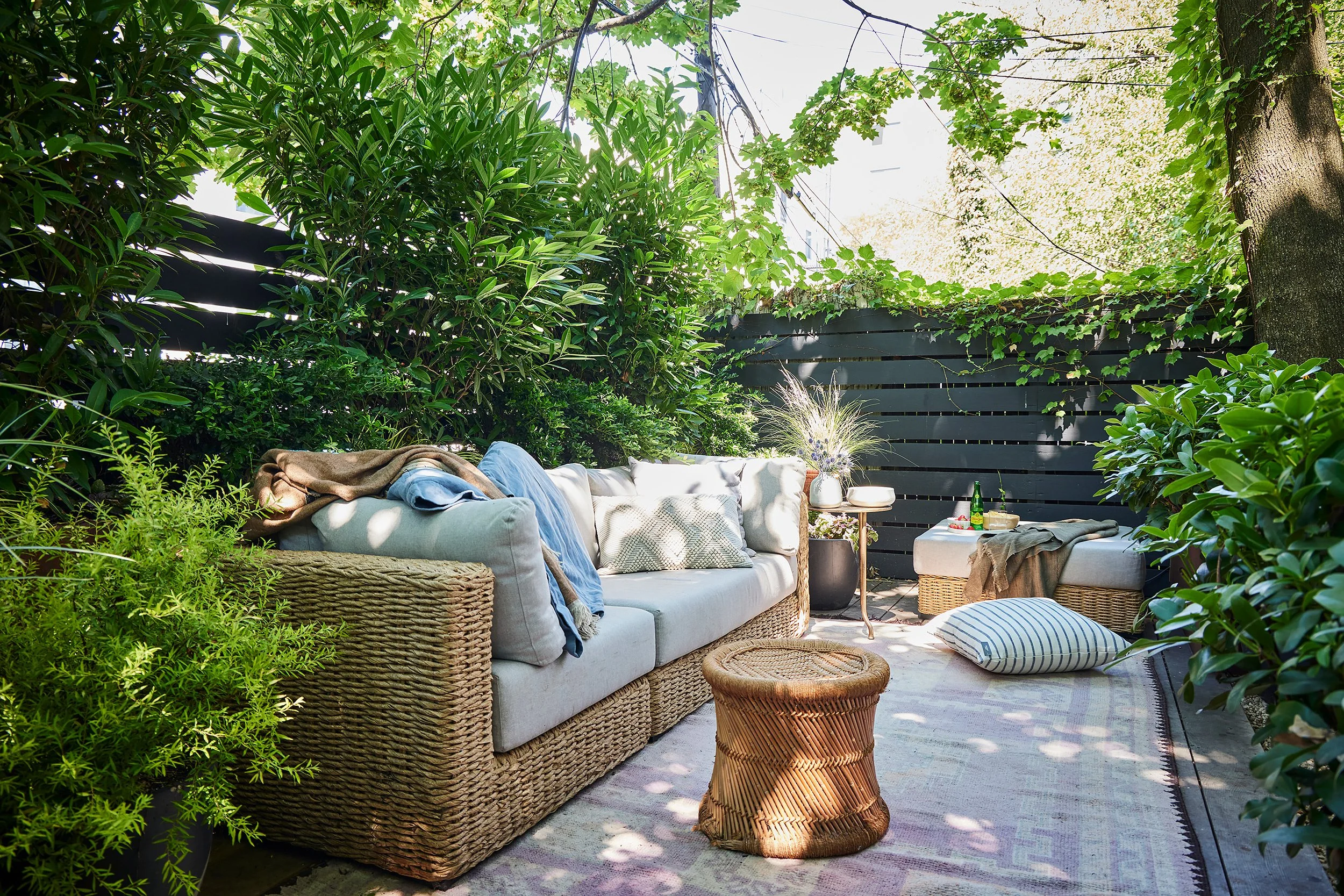 Outdoor patio with a wicker sofa topped with cushions and throws, a small round wicker coffee table, falling sunlight, lush green plants, potted flowers, a striped pillow on the ground, and a dark fence in the background.