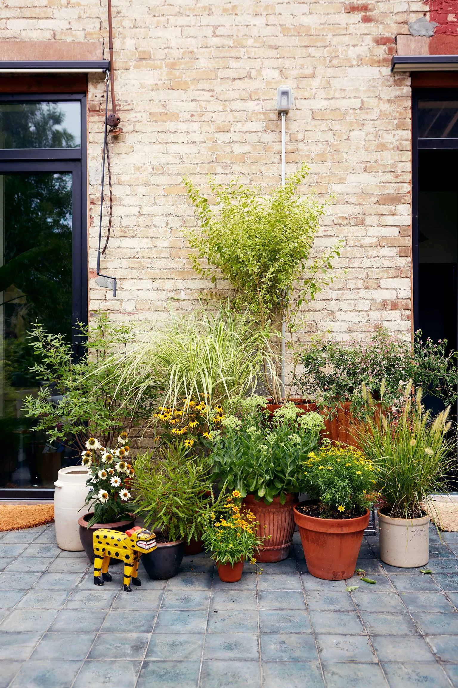Container garden with various potted plants and flowers against a brick wall, including a yellow and black decorative tiger figure.