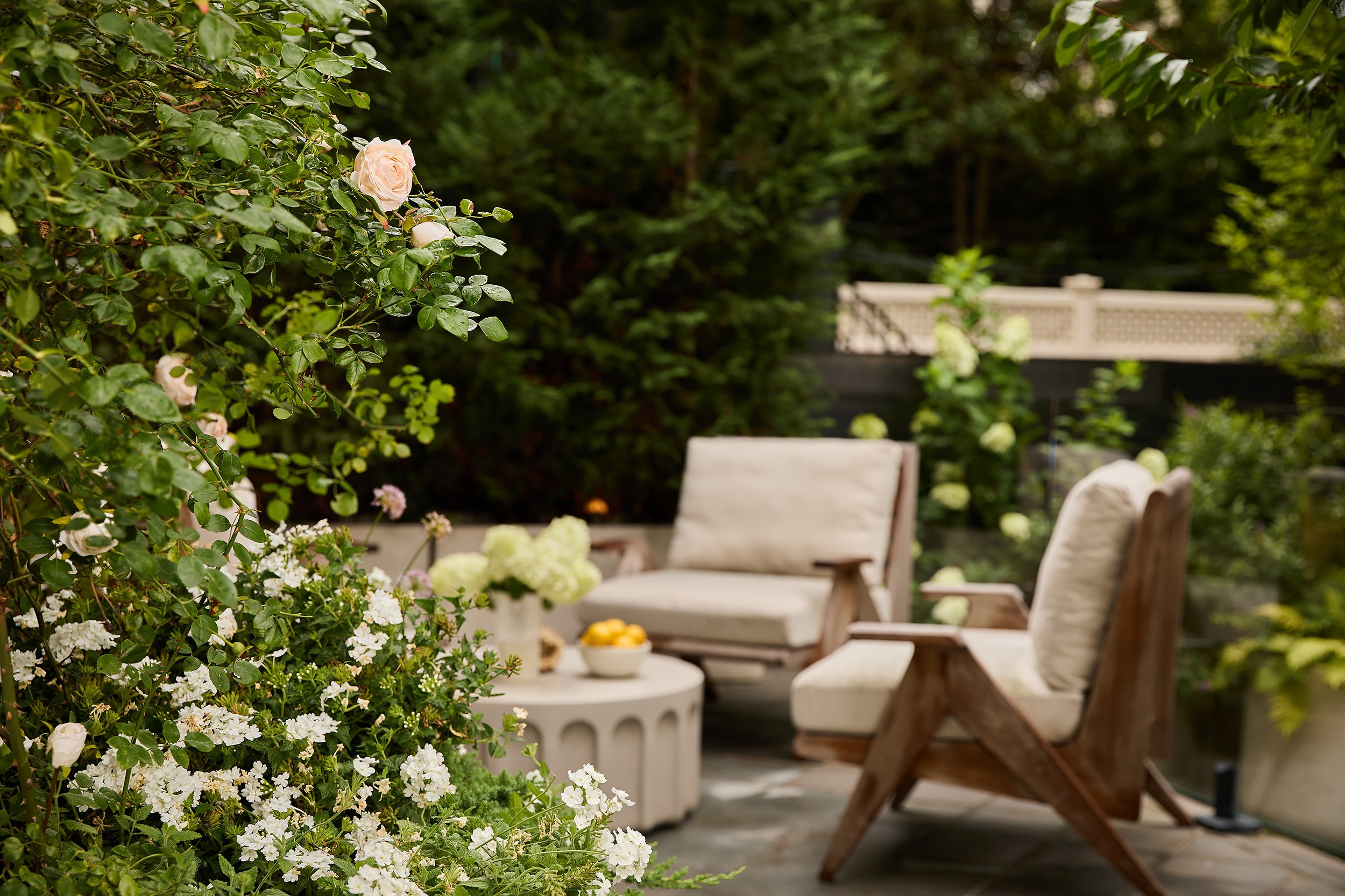 A cozy outdoor patio with two cushioned lounge chairs, a small white table with a bowl of lemons, surrounded by lush green foliage and blooming flowers.