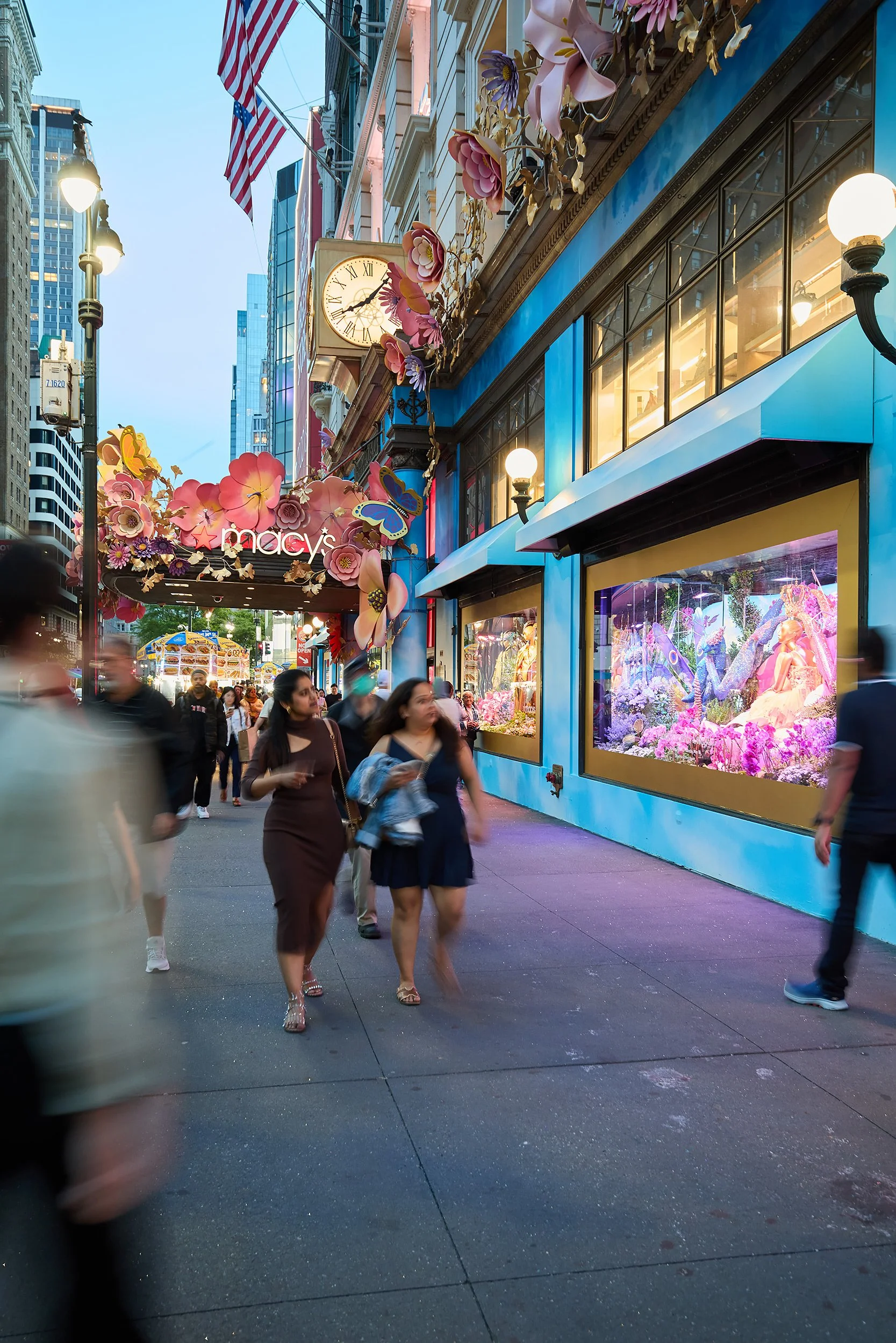 Pedestrians walking past a Macy's store decorated with pink floral and butterfly ornaments on a city street in the evening.