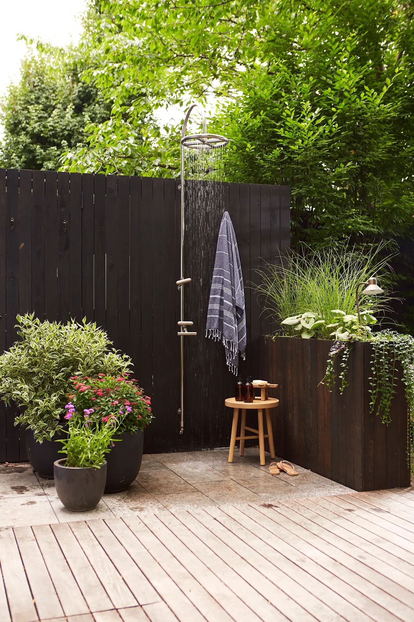 An outdoor shower area with a black wooden privacy fence, surrounded by potted plants and greenery. There is a round wooden stool with bottles on top, a pair of sandals on the wooden deck, and a towel hanging from the shower. The scene is lush and peaceful.