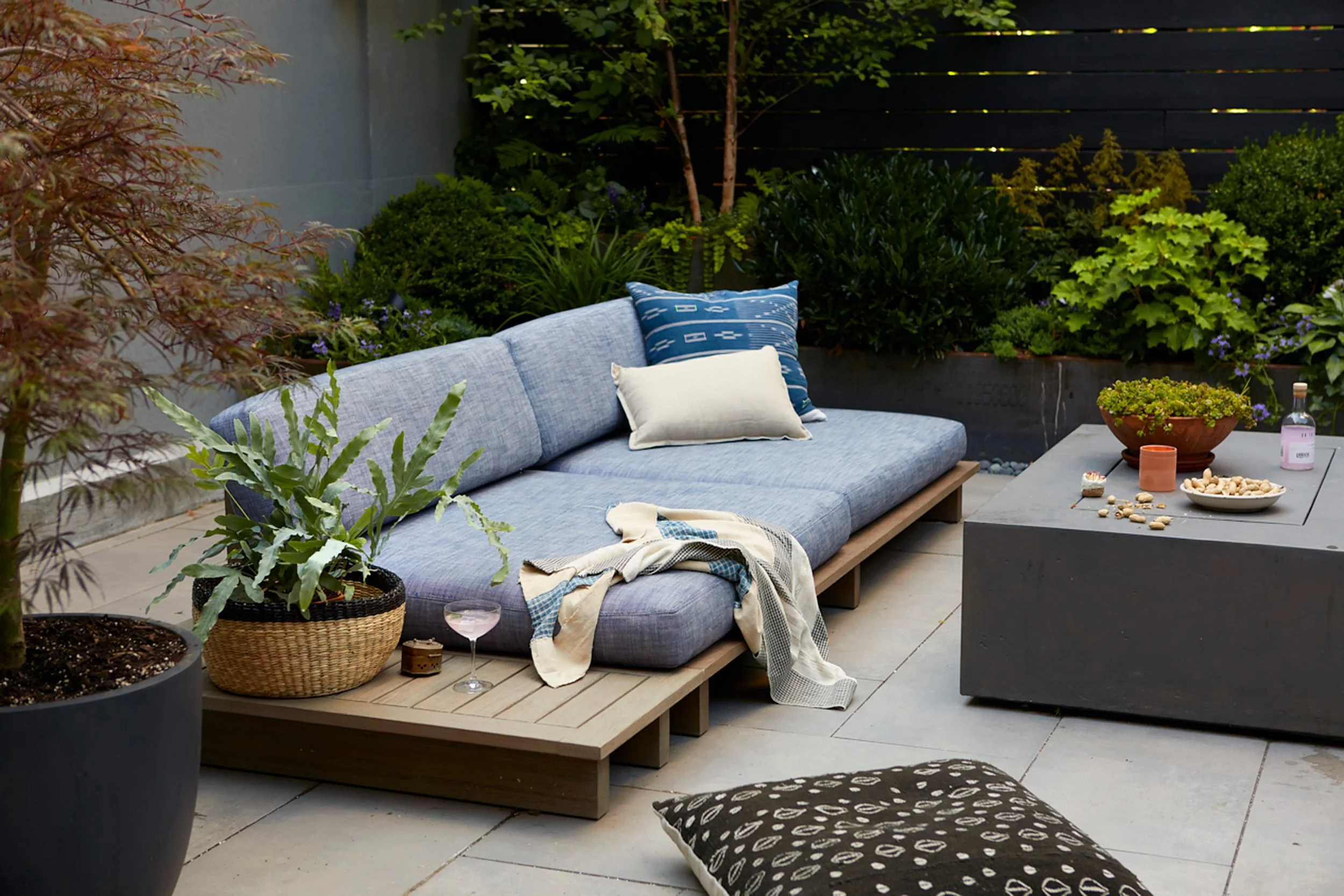 Outdoor patio with a blue cushioned sofa, coffee table with snacks, and surrounding greenery.