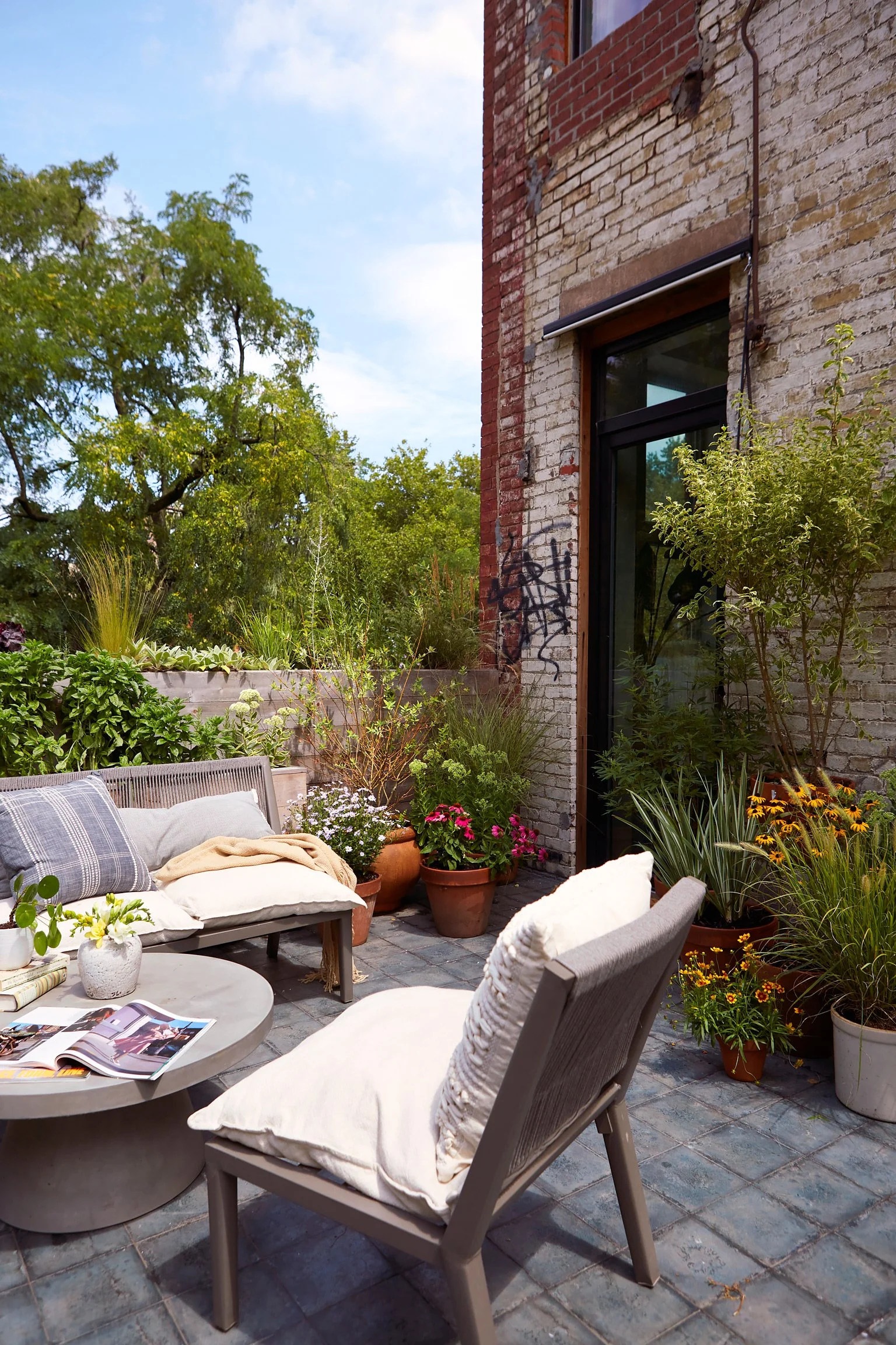 Outdoor patio space with beige cushioned chairs, potted plants, and greenery against a brick building with graffiti and large windows.