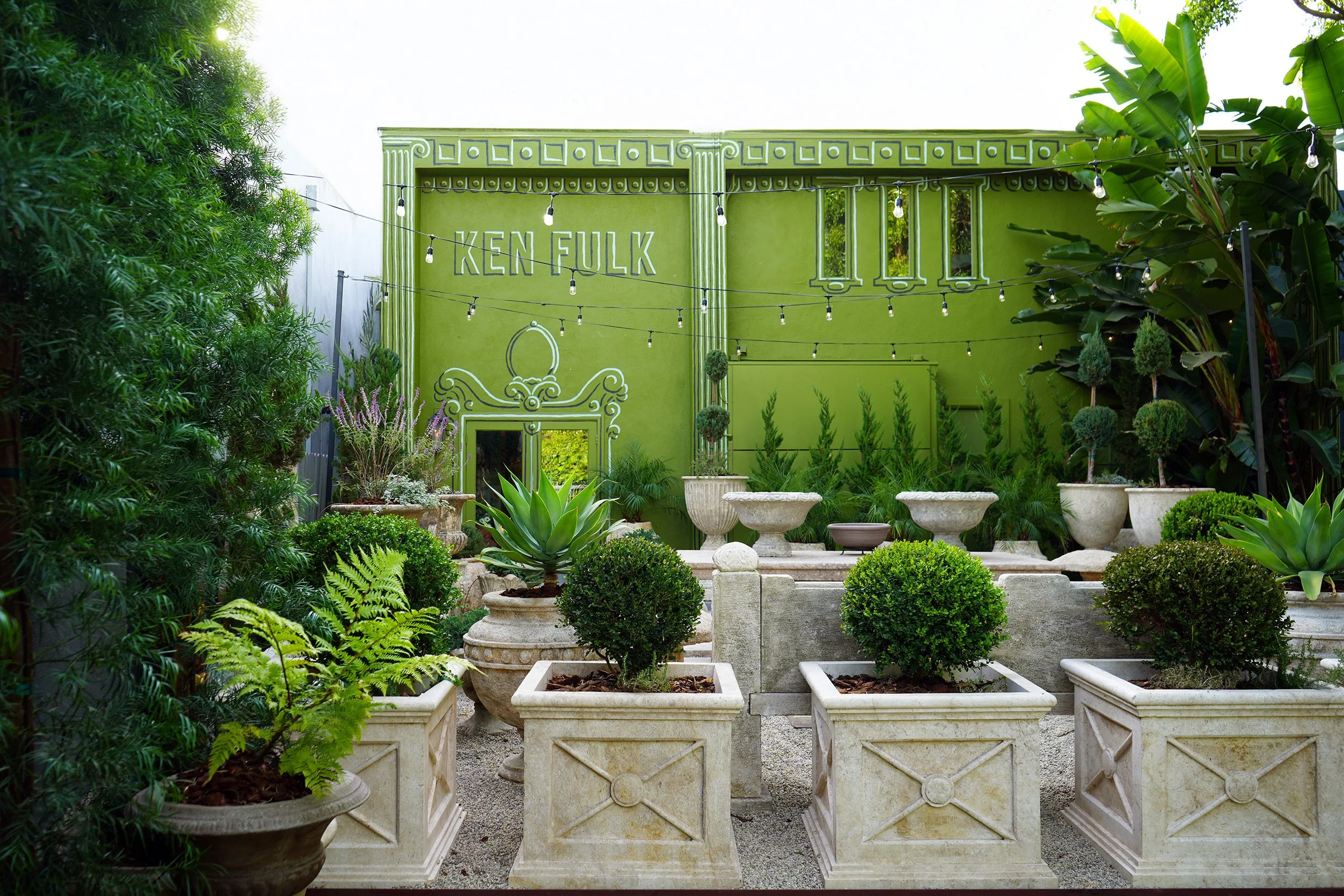 A lush garden space with vibrant green walls, potted plants, and decorative urns, featuring a sign that says 'Ken Fulk'.