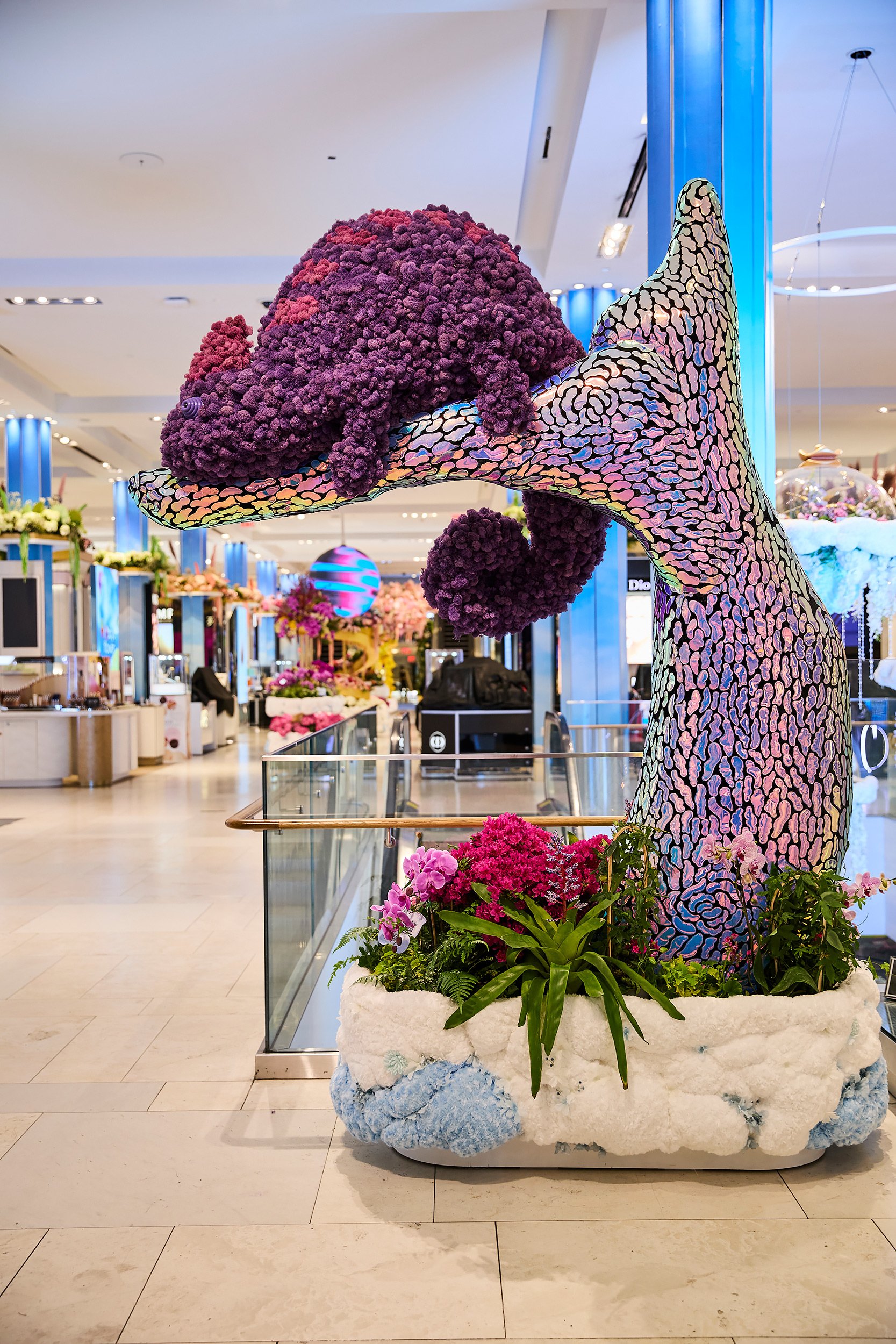 Artificial sculpture of a chameleon made of colorful mosaic tiles, with a body composed of purple flowers, situated in a shopping mall decorated with other floral displays.