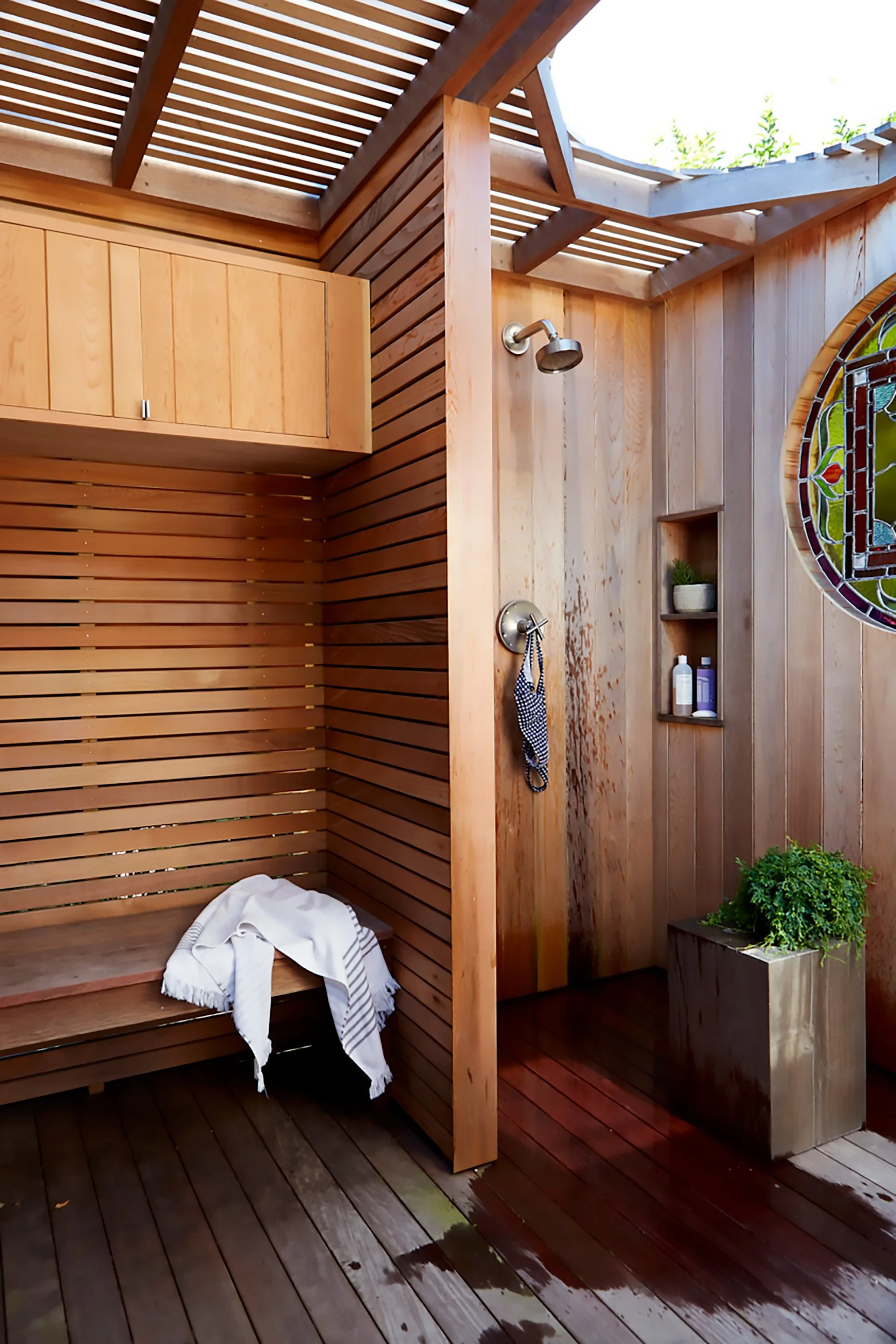 Outdoor wooden shower with a bench, hanging towel, small shelves with toiletries, and a stained glass window.