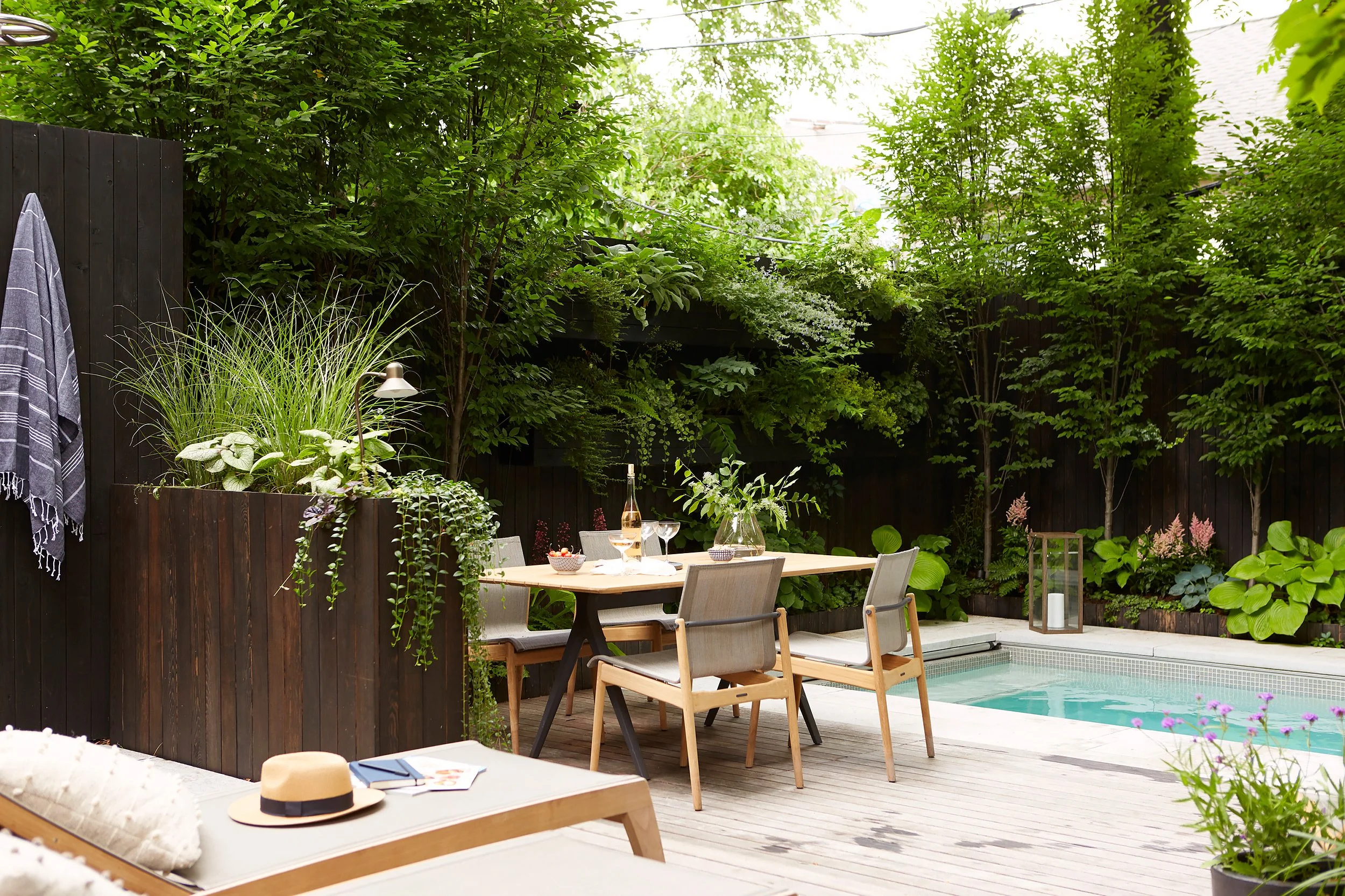 A backyard patio with a small swimming pool, a dining table set with chairs, and lush green trees and plants surrounding the area.