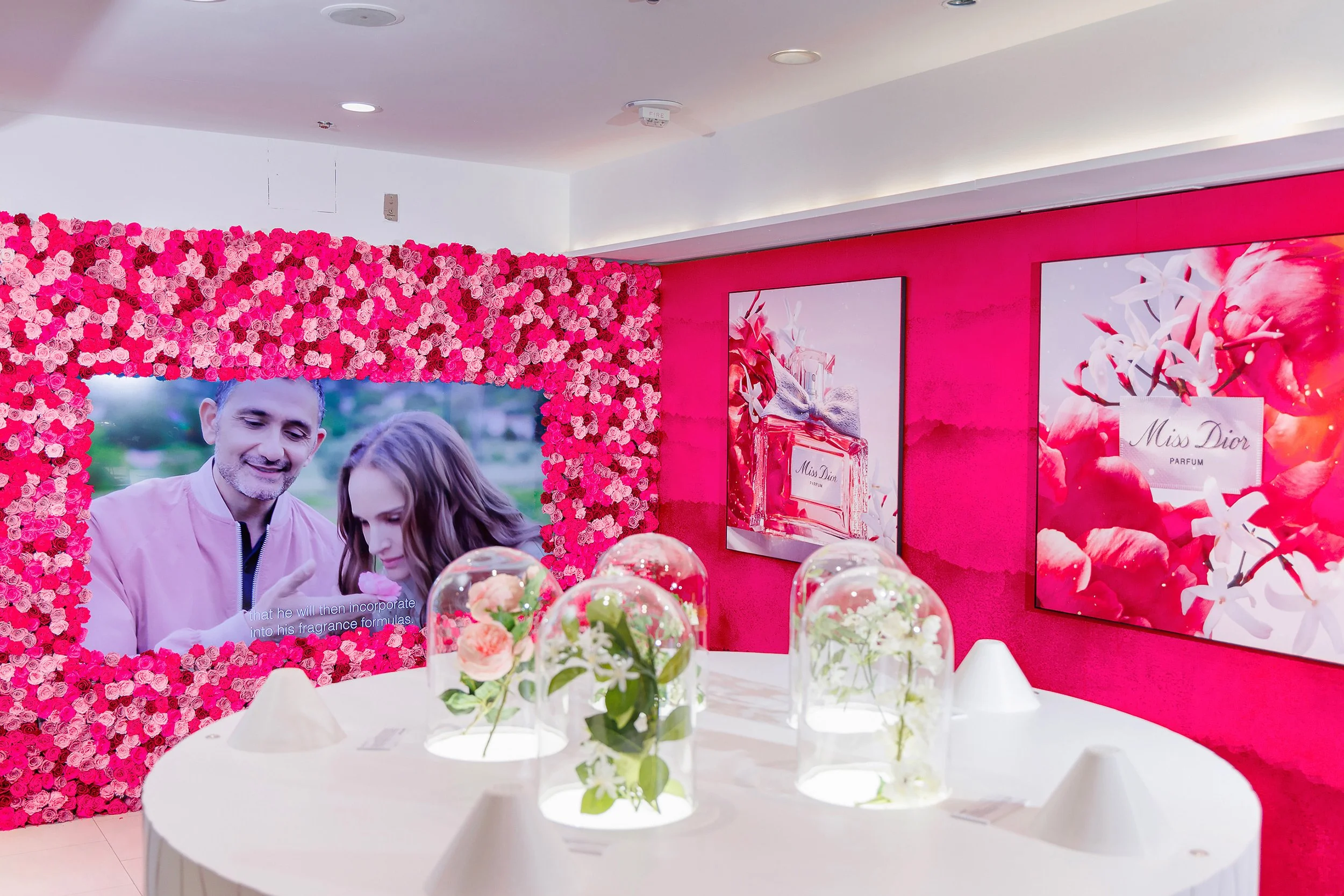 A display area with a large digital screen surrounded by a wall of pink roses, showing a couple. To the right, pink floral-themed advertisements for Miss Dior perfume are on the wall. On a white table in front, several glass domes containing flowers are arranged.