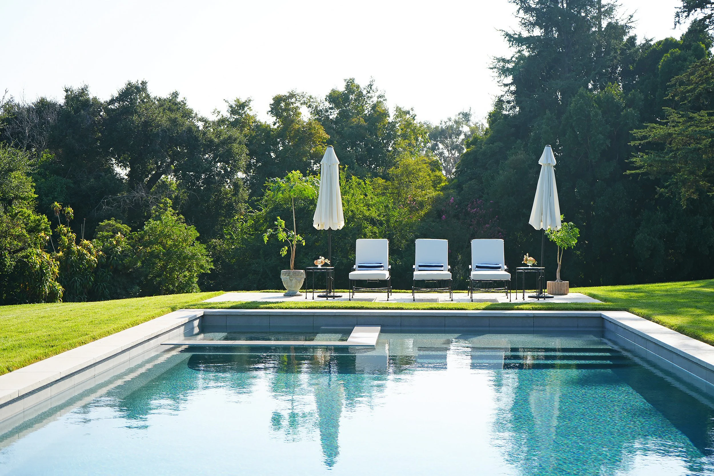 Swimming pool with four white lounge chairs and two umbrellas, surrounded by greenery and trees.