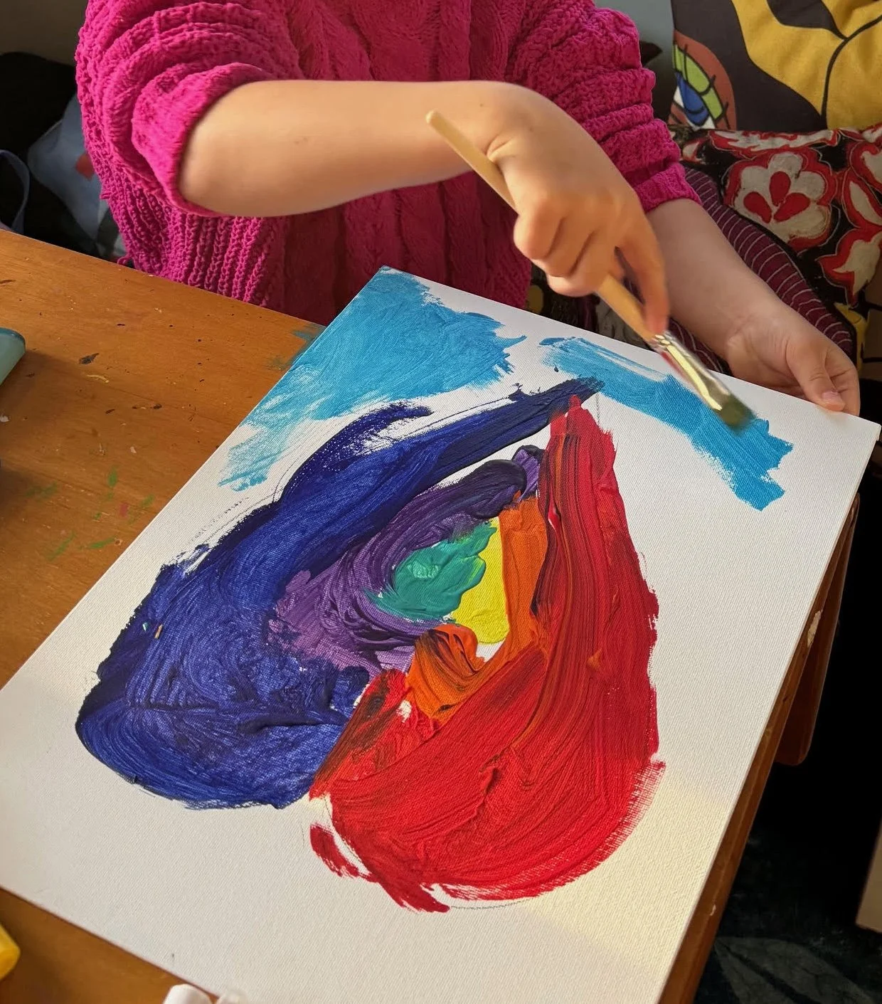 An image of a child's hands holding a paintbrush adding intense blue to a canvas of a painted heart in rainbow colors