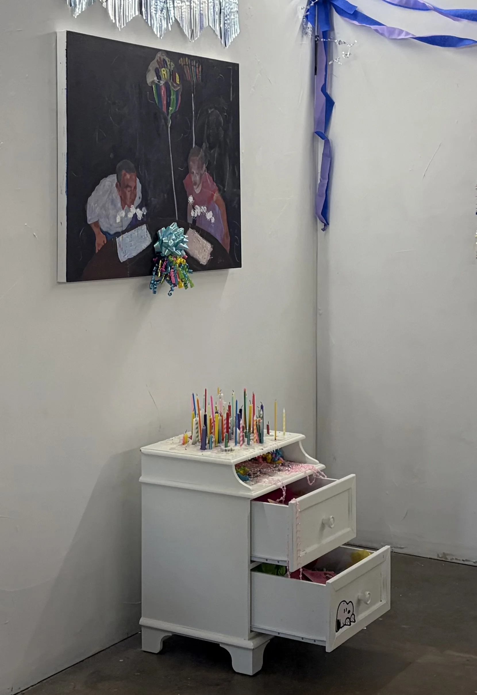 A detail of an art installation including a painting of two people blowing out birthday candles hanging on the wall above a white two drawer side table with candles and confetti on its surface