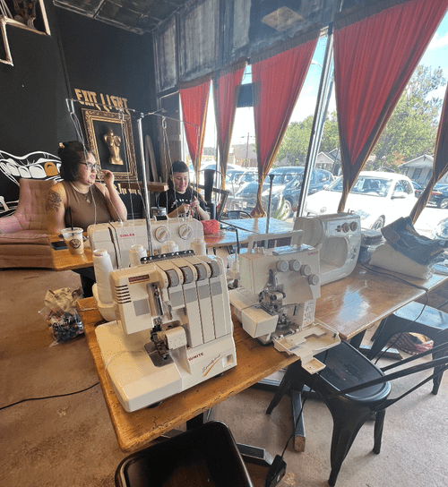 Two people sit in a coffee shop with black painted walls and gold graphic murals, working on knitting and embroidery projects in front of tables full of sewing machines and sergers