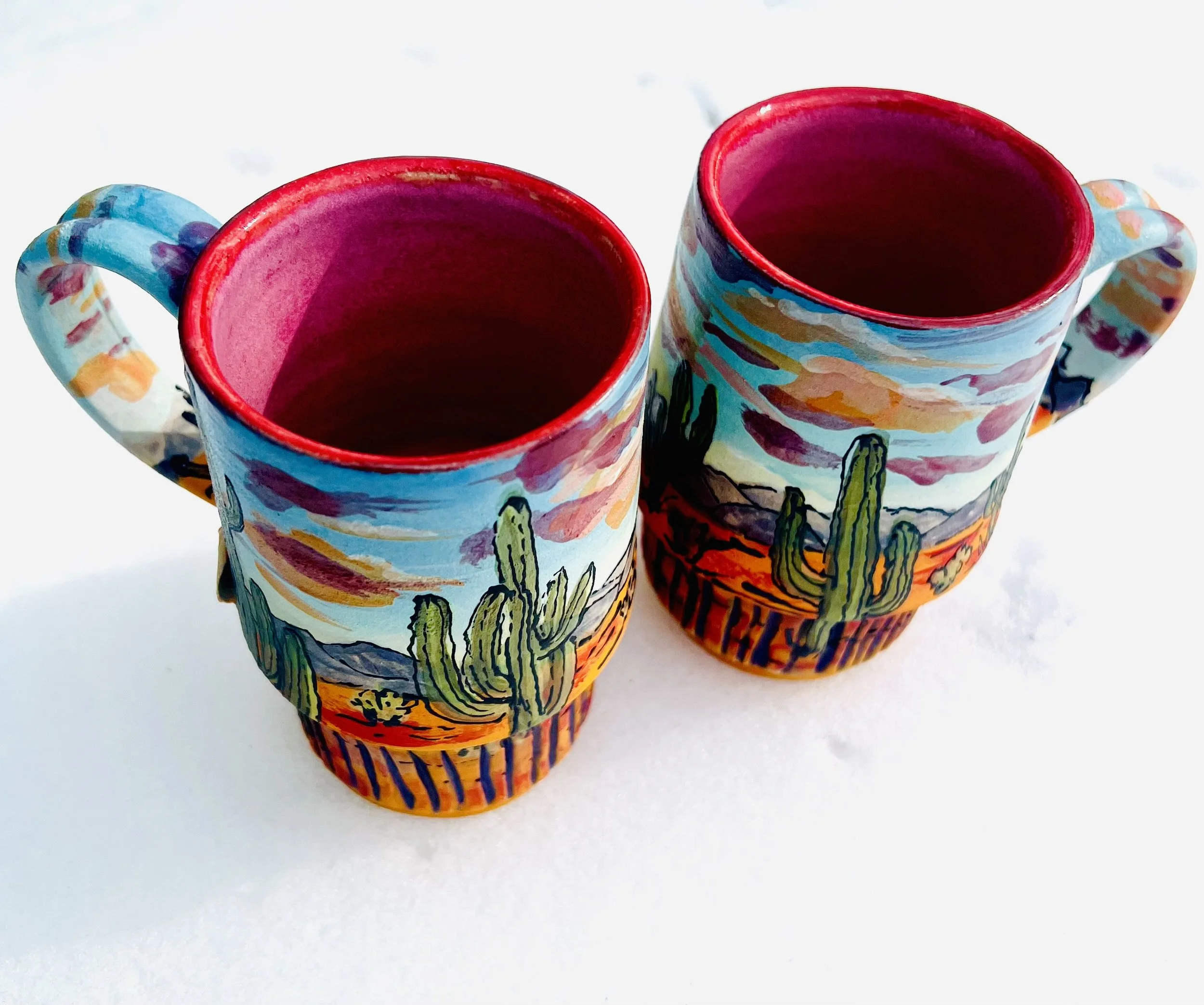 Two ceramic mugs painted bright red inside with a desert landscape of cacti and mountains agains a sunset painted on the outside