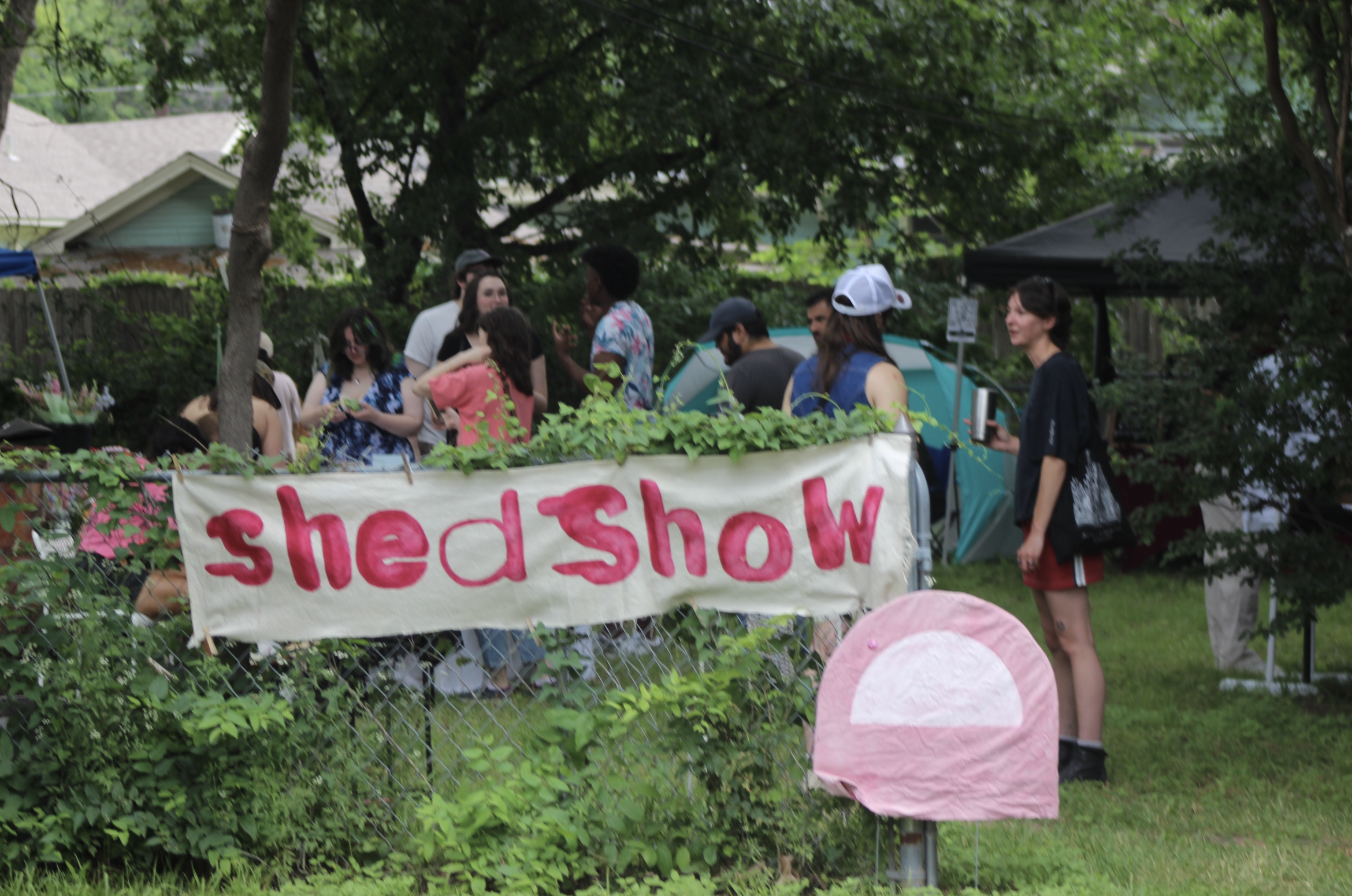 A backyard full of people behind a fence holding a banner with 'shed show' hand painted in red, drippy letters