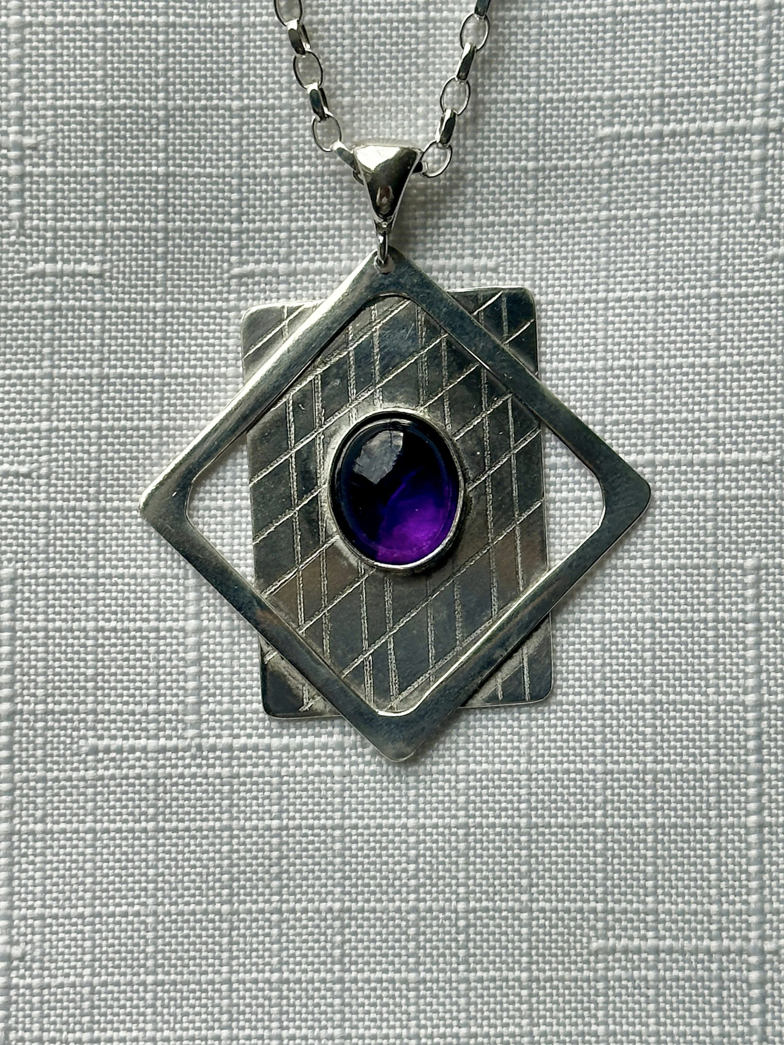 A minimalist silver rectangle pendant with perpendicular lines etched in the surface and a deep purple stone set in the center