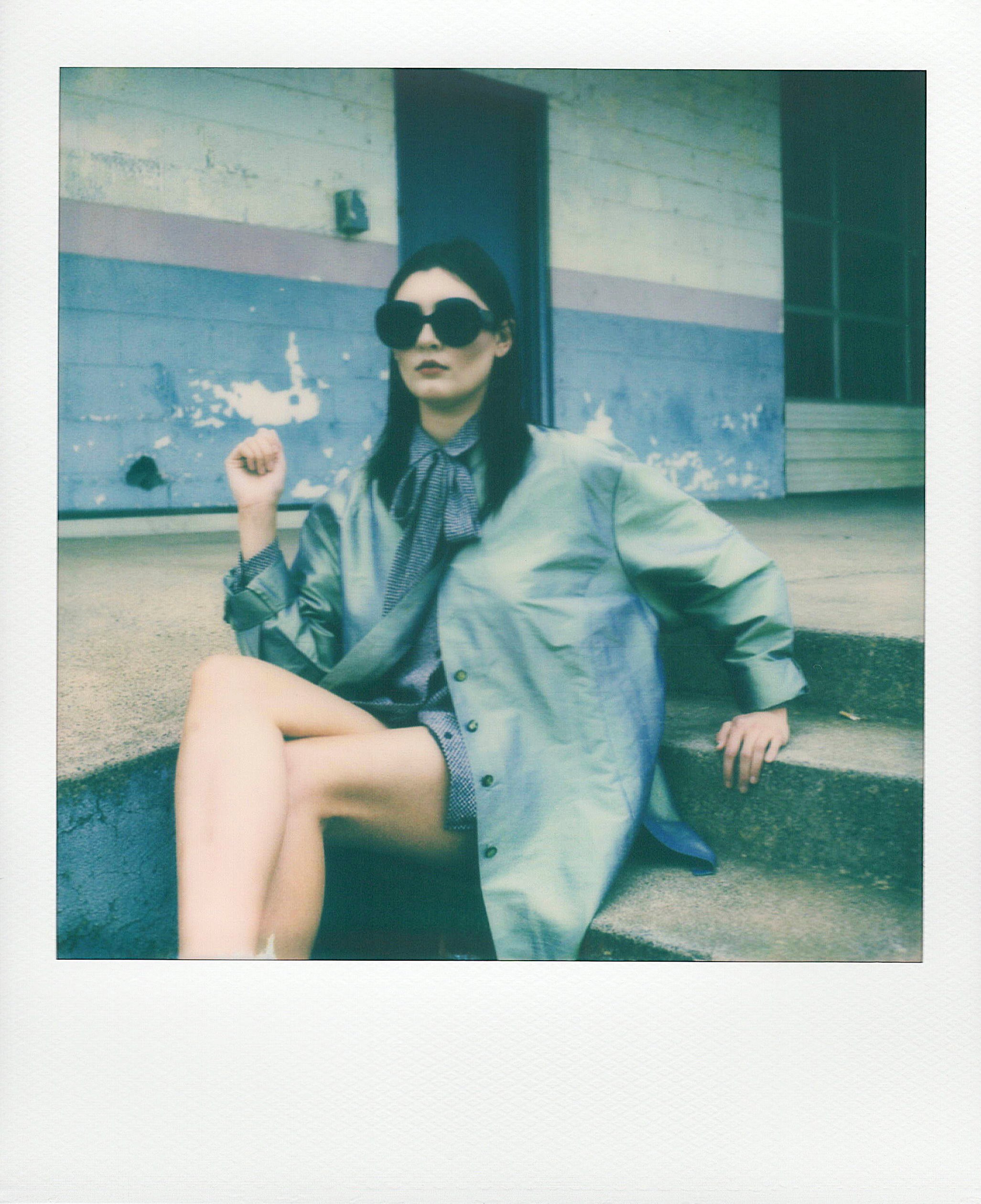 A polaroid of a femme person with long dark hear and large dark sunglasses wearing a blue overshirt and a blue dress with a bow at the neck sitting on cement steps
