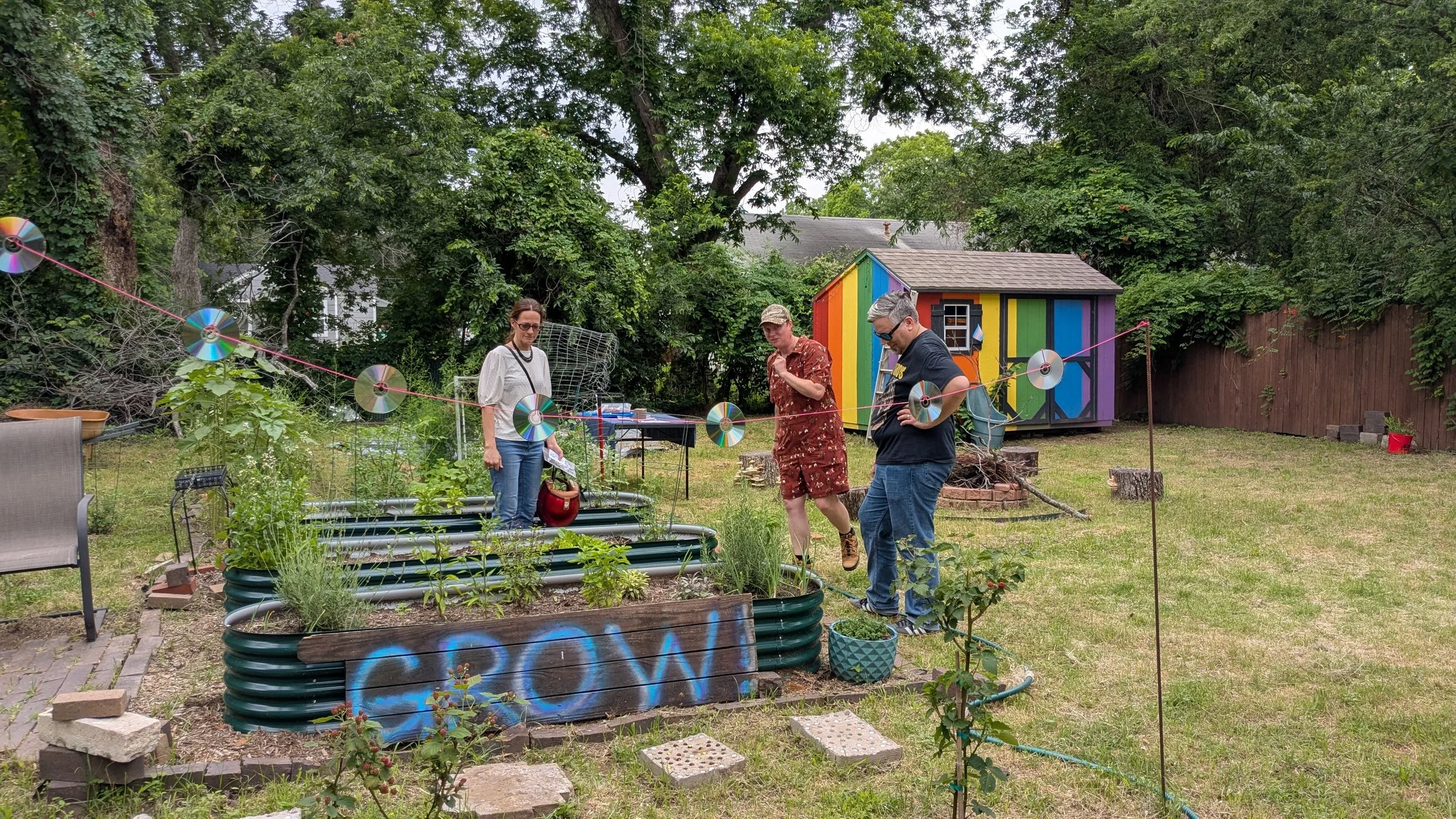 Three people in a backyard garden with a painted sign that says GROW! and a rainbow painted shed in the back