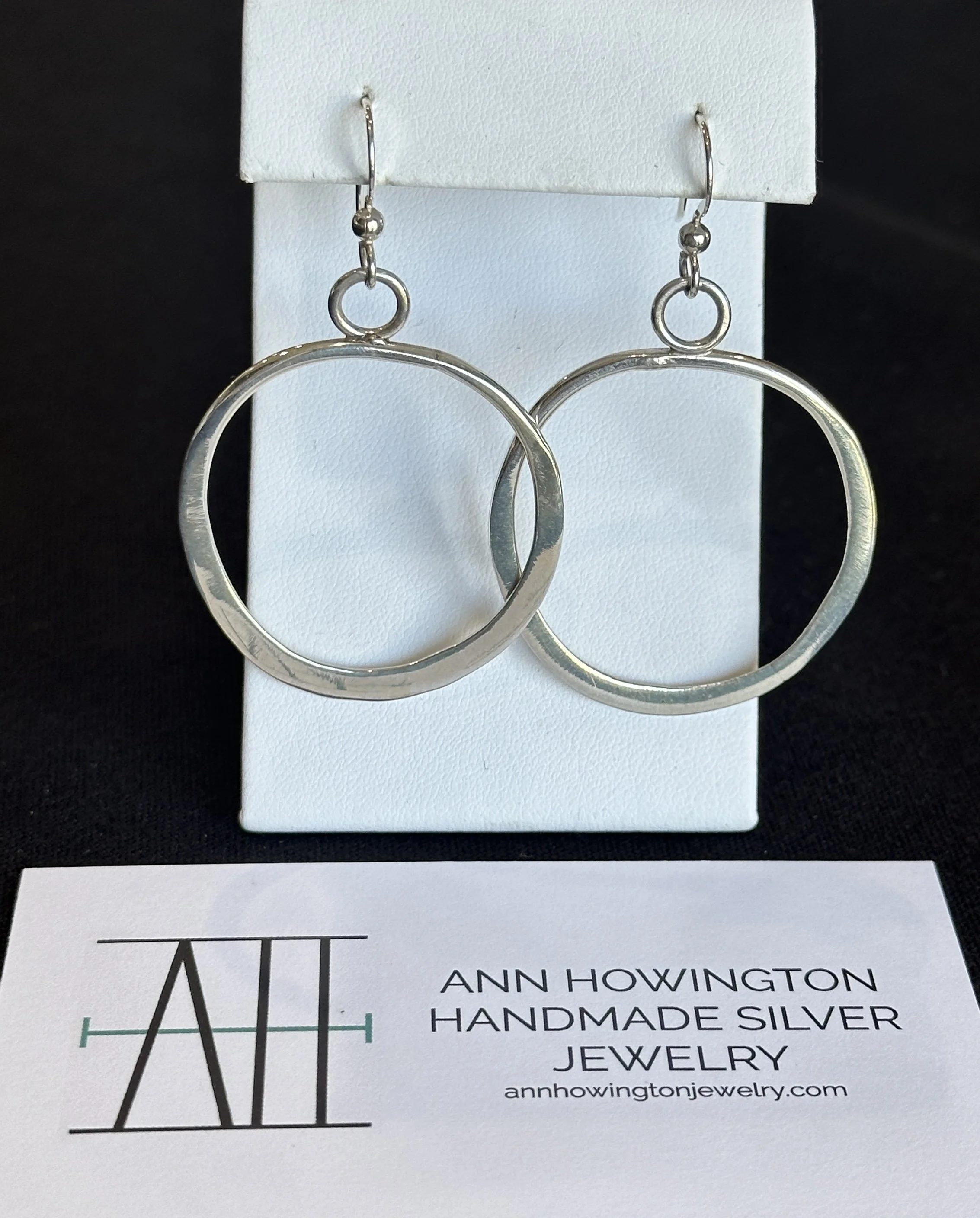 A pair of silver circular earrings in front of a Ann Howington's business card
