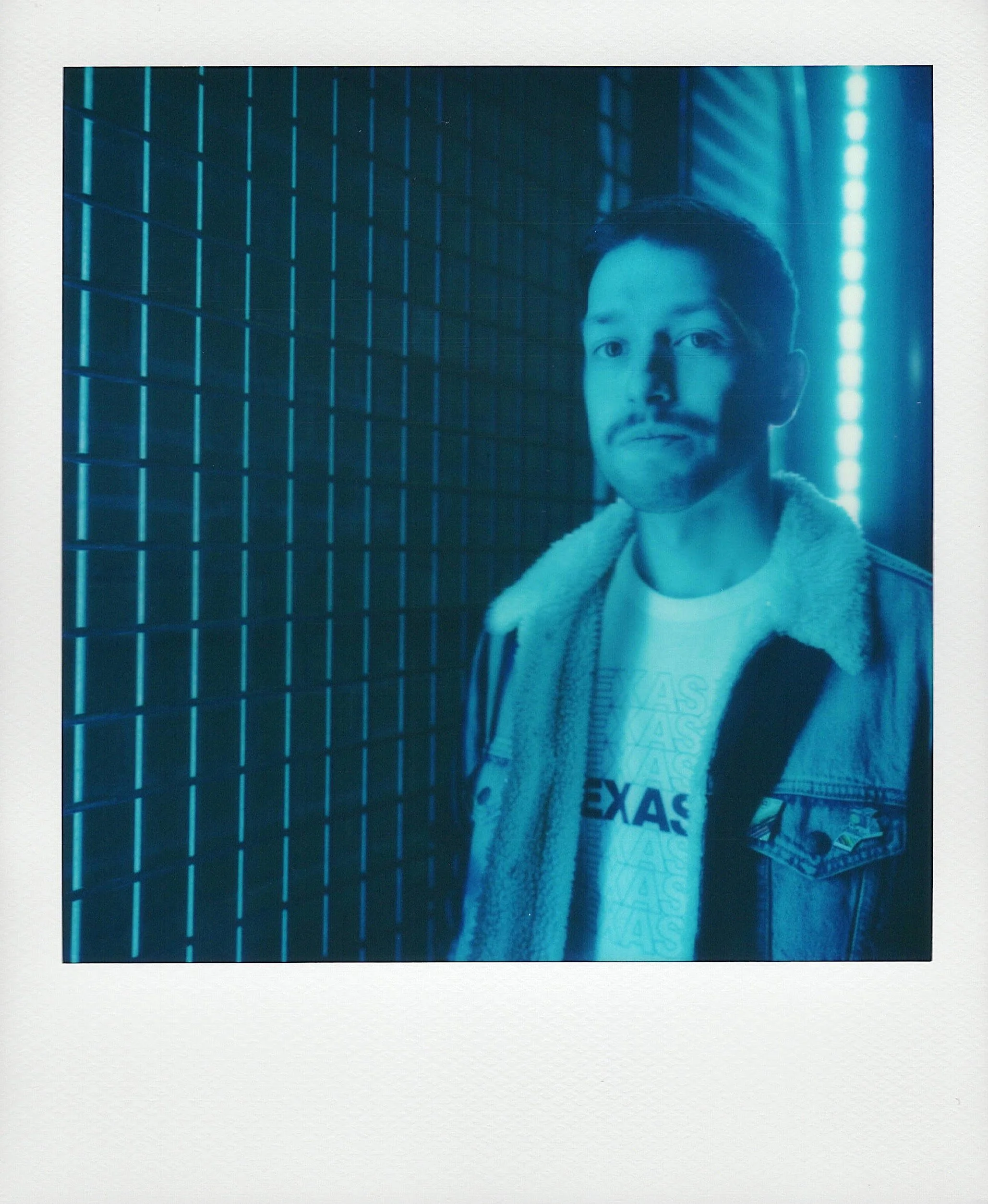 A polaroid with a blue color cast showing John Eric Munoz looking at the camera wearing a denim jacket and a tshirt that says Texas