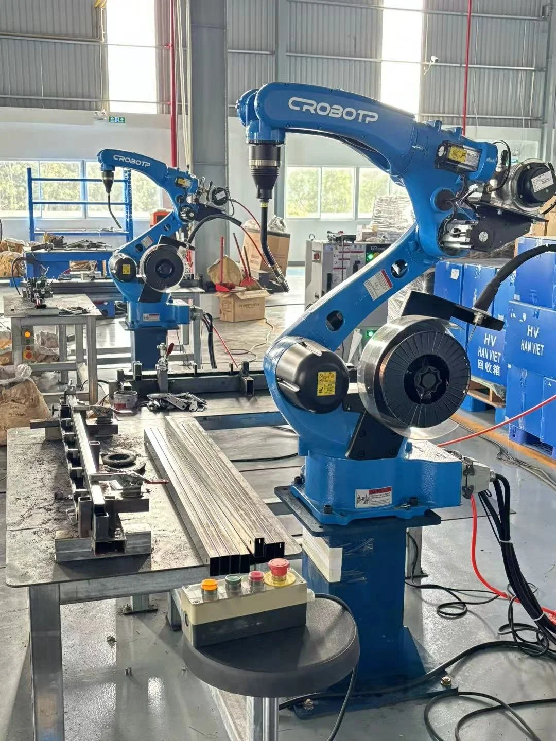 Two blue industrial robotic arms working in a manufacturing facility, with one arm holding an object near a worktable.