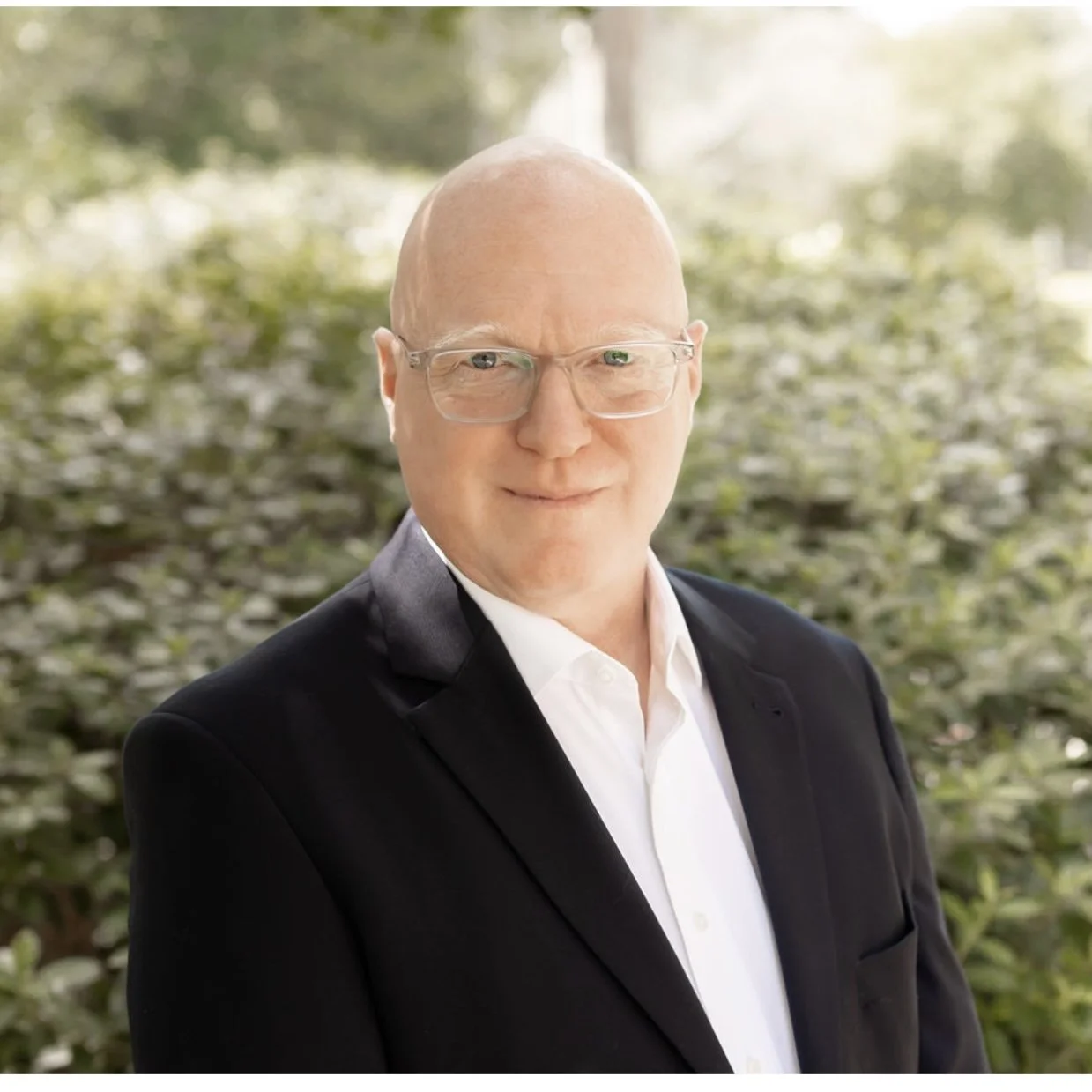 Portrait of a middle-aged man with glasses, dressed in a black suit and white shirt, standing outdoors with blurred green foliage in the background.