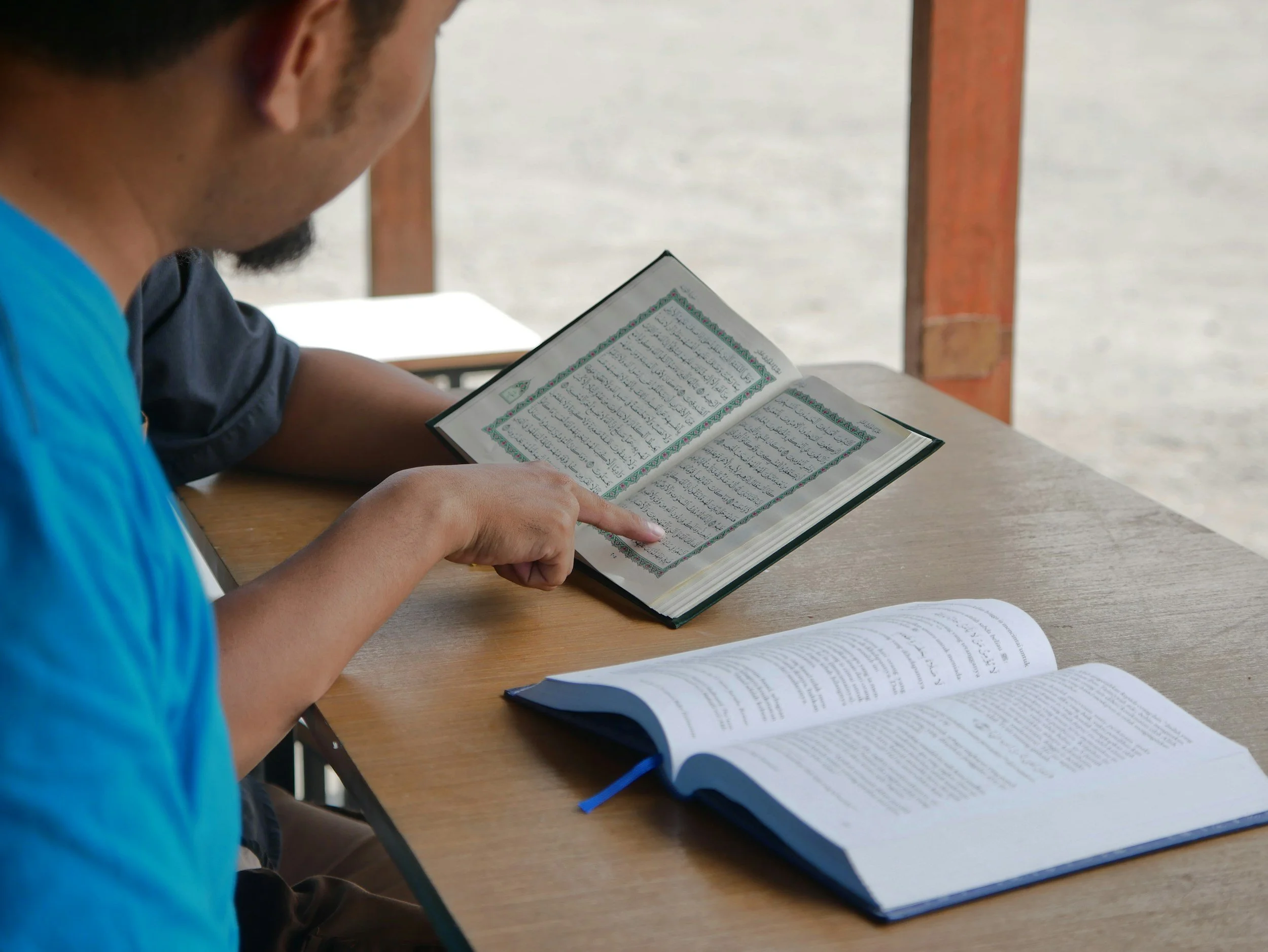 Un hombre con camiseta azul lee un libro que contiene texto en árabe, sentado en una mesa de madera con otros libros abiertos frente a él.