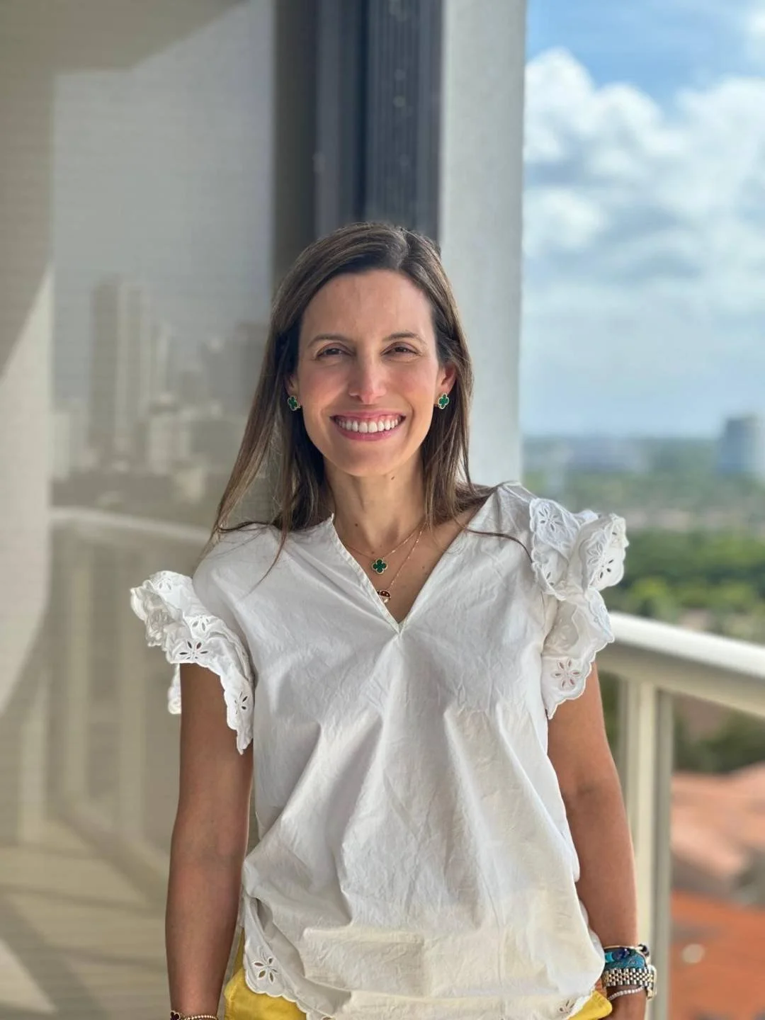 Mujer sonriendo con blusa blanca de manga corta con detalles de encaje, grandes pendientes de trébol, reloj y pulsera. Fondo de ciudad y cielo nublado visto desde un balcón.