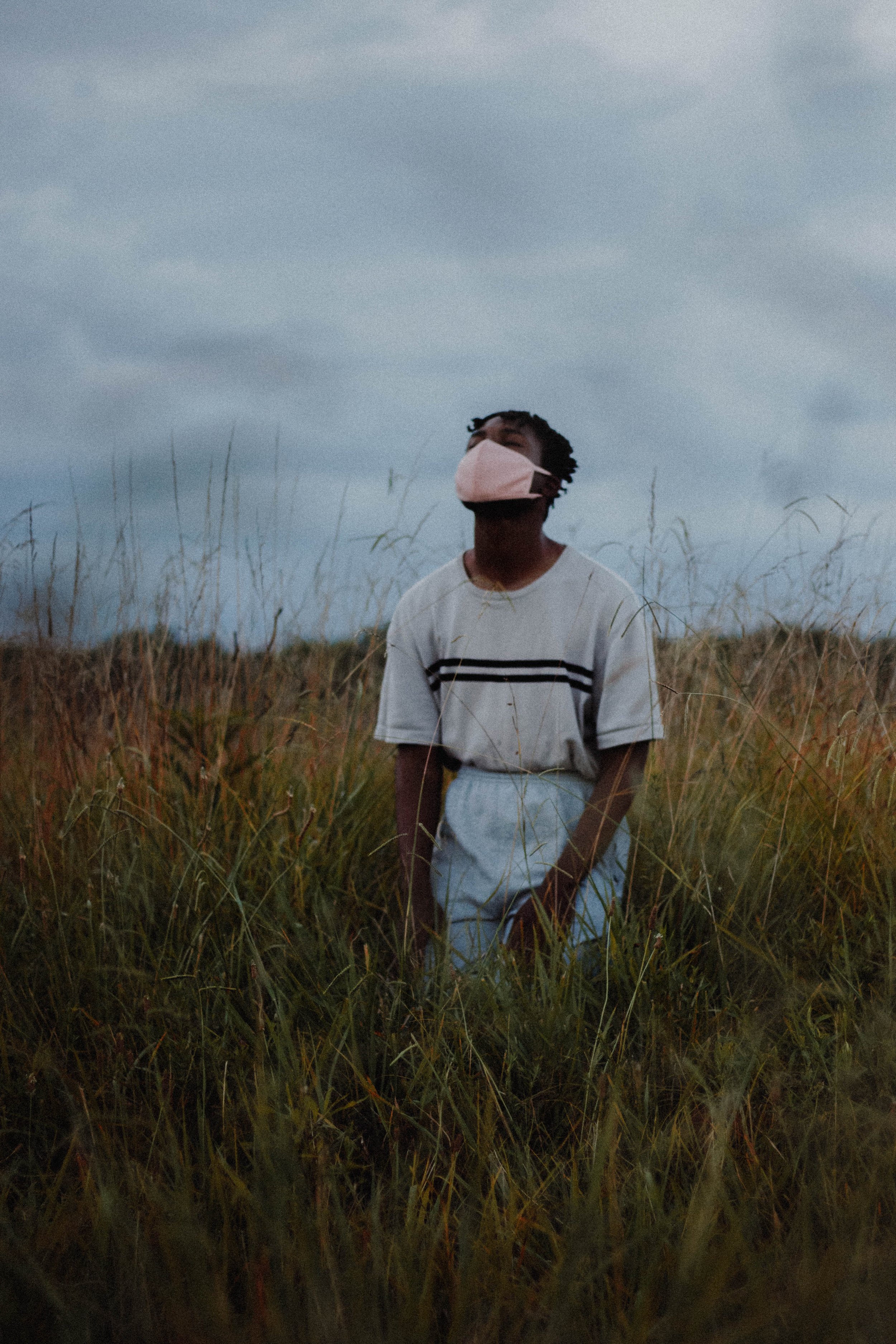 A person wearing a face mask and a casual outfit standing in tall grass outdoors under a cloudy sky.