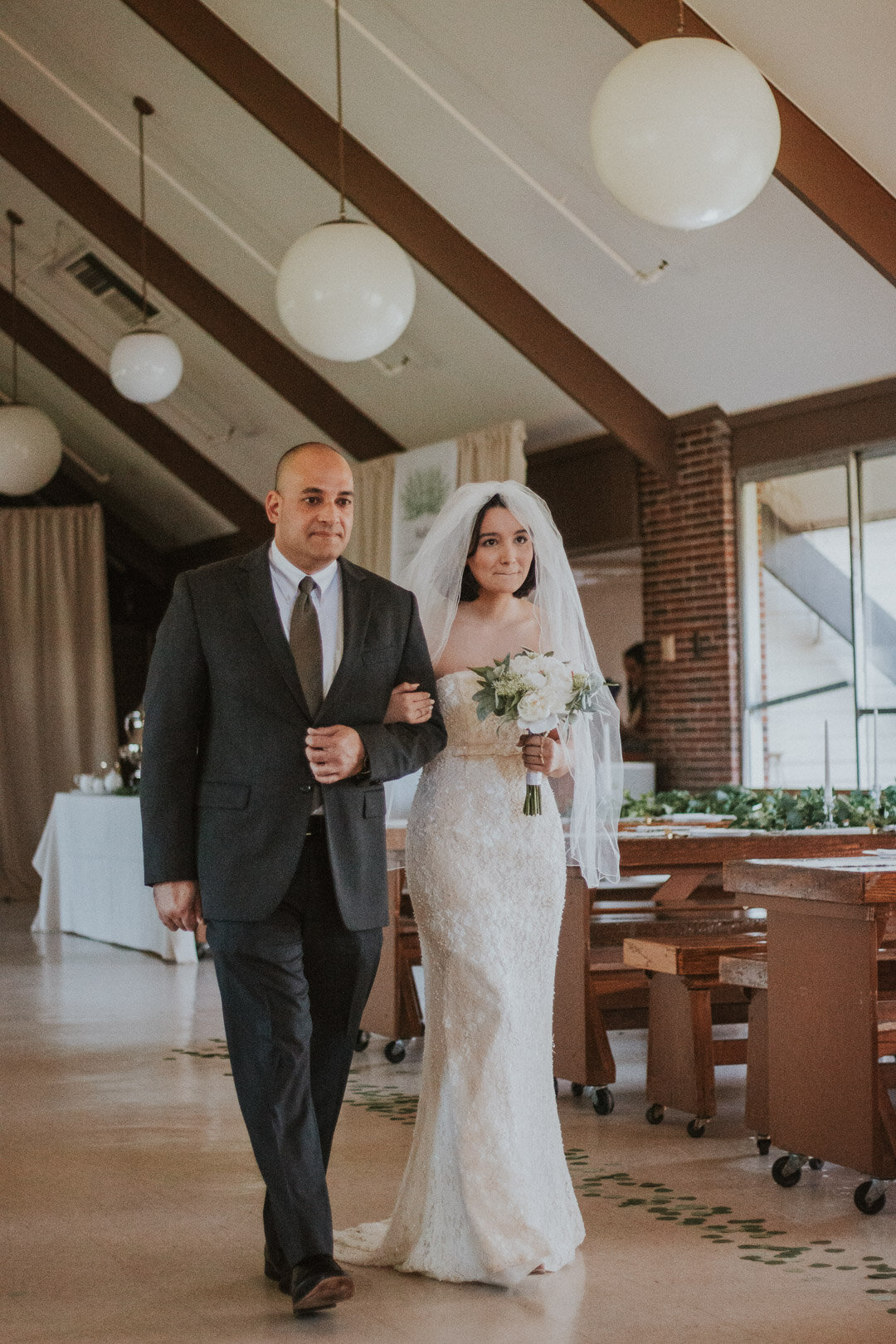 Bride in a white lace wedding gown and veil holding a bouquet of white flowers, walking down the aisle with a man in a dark suit with a white shirt and tie inside a decorated reception hall.