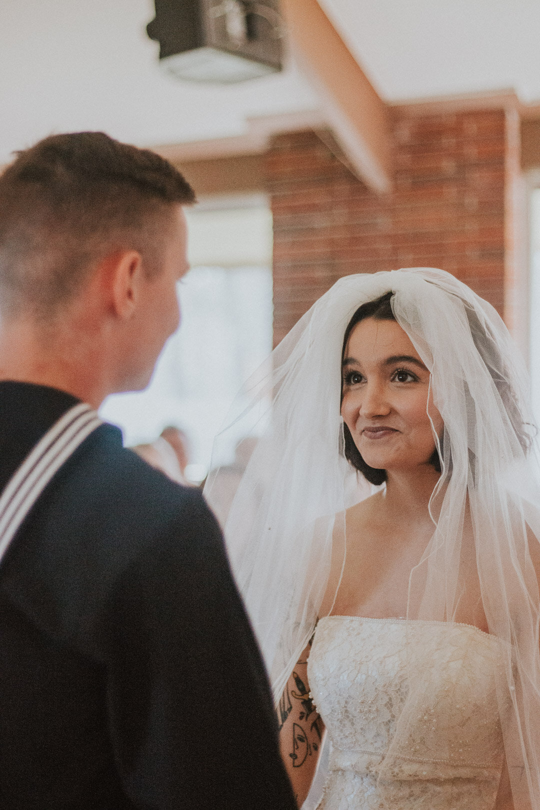 A bride with dark hair, wearing a strapless lace wedding dress and a veil, looks at a groom with short brown hair, dressed in a dark suit, during their wedding ceremony indoors.