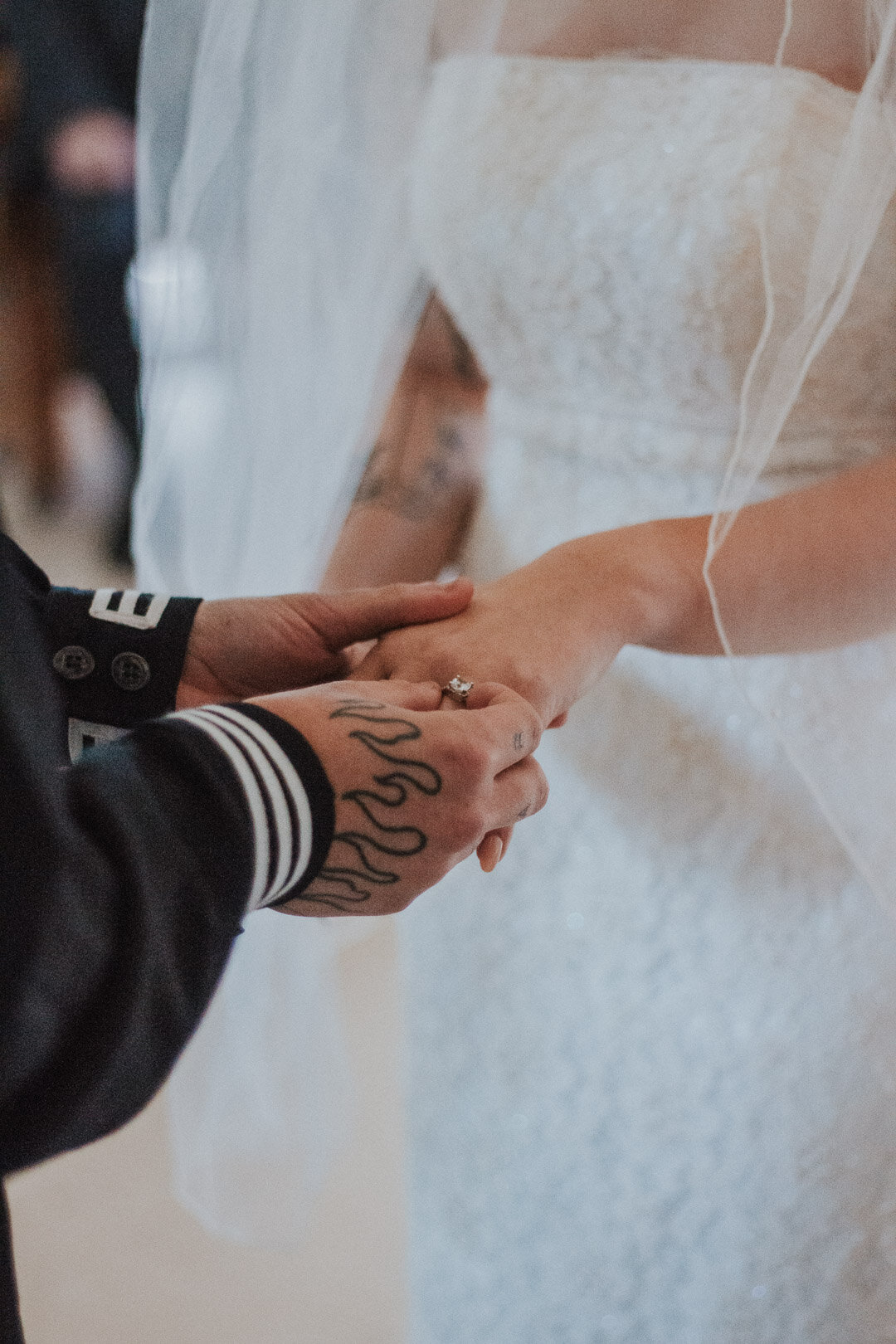 Close-up of a couple holding hands during a wedding ceremony, with the bride in a white dress and veil, and the groom wearing a dark jacket with tattooed hands.