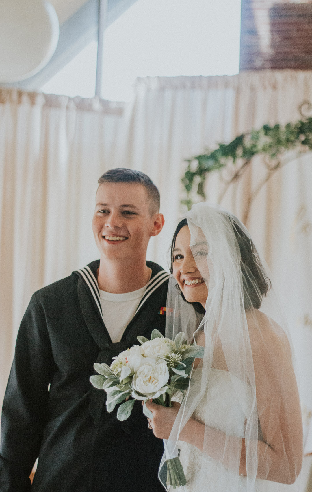 A bride and groom smiling together at their wedding, with the bride holding a bouquet of white flowers, and a light-colored curtain and greenery in the background.